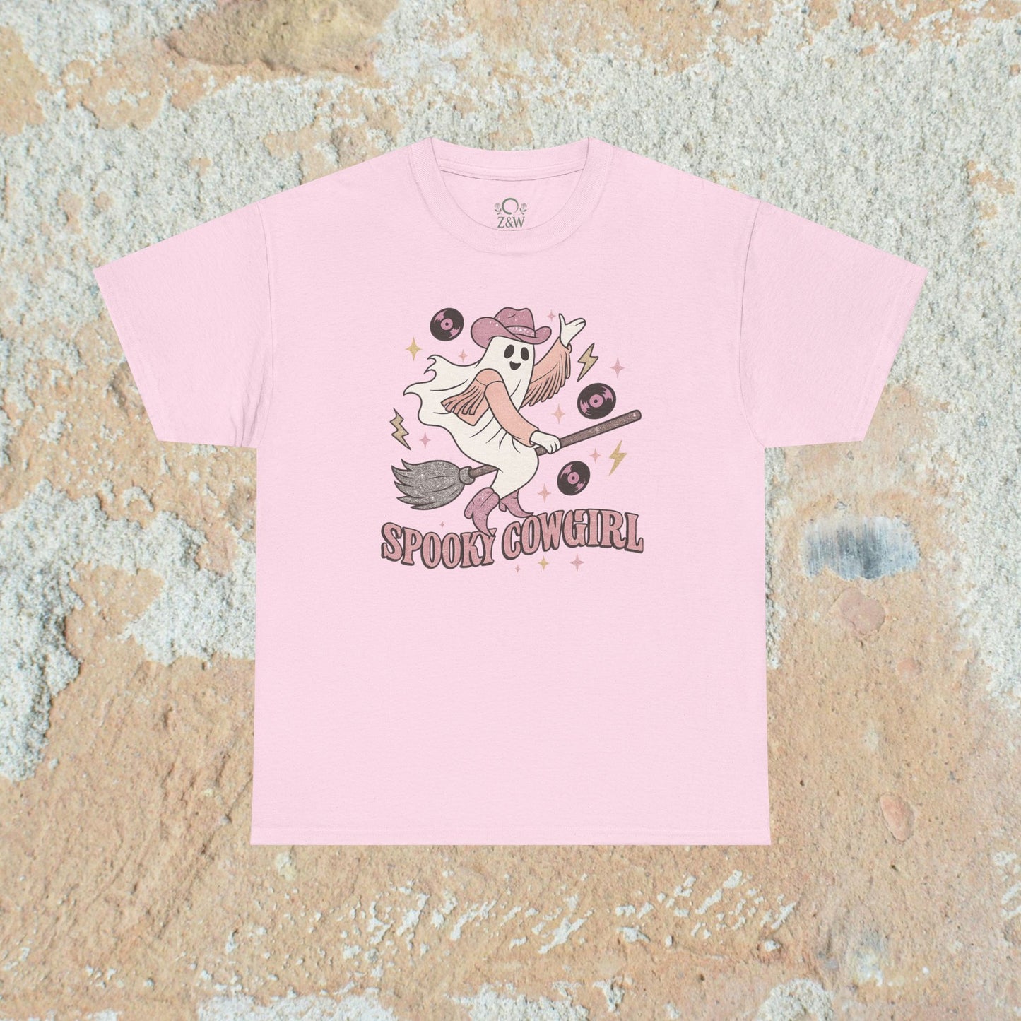 Spooky Cowgirl Graphic Tee in soft light pink showcasing a whimsical ghost character with a hat and broom, ideal for festive occasions and everyday fun.