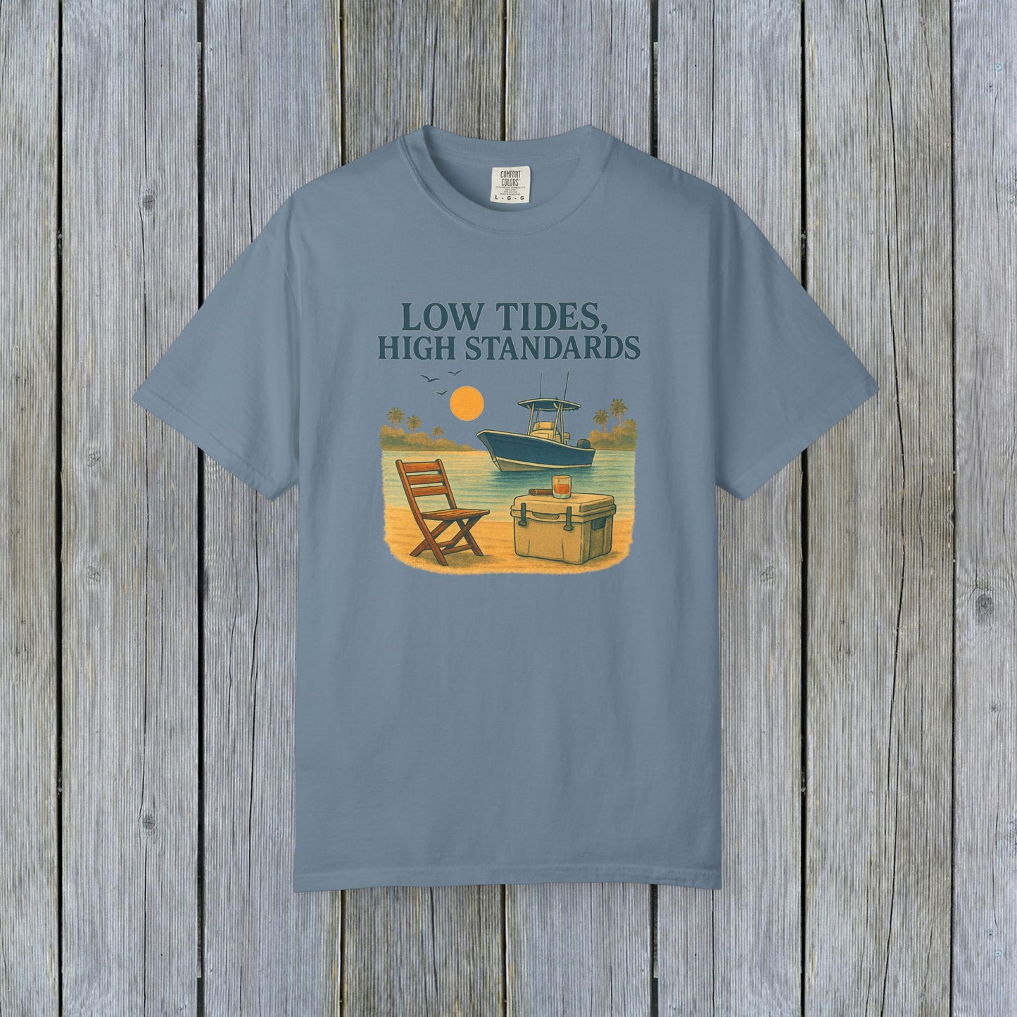 Garment-Dyed Low Tides High Standards T-Shirt in blue color, showcasing a vibrant beach illustration with a chair and cooler beside a boat. This soft 100% US cotton tee is offered in sizes S-4XL.