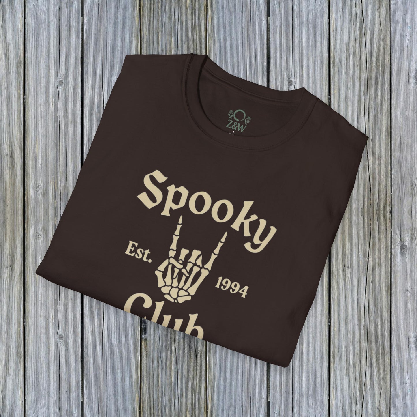 Folded Unisex Spooky Club Tee in dark brown, placed on a wooden backdrop, featuring spooky skeleton hand artwork and 'Spooky Club Est. 1994' tagline. Soft 100% cotton fabric perfect for Halloween.