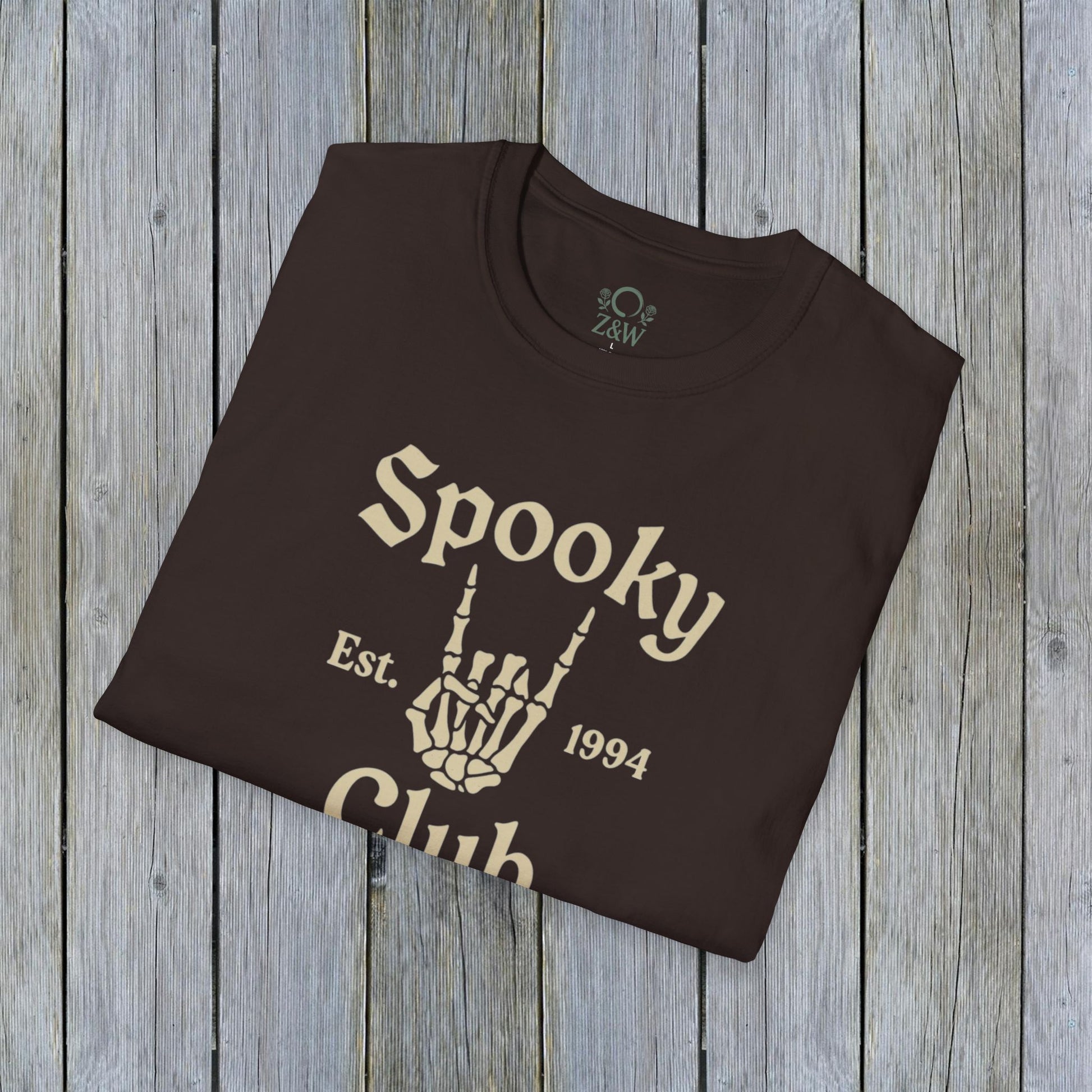 Folded Unisex Spooky Club Tee in dark brown, placed on a wooden backdrop, featuring spooky skeleton hand artwork and 'Spooky Club Est. 1994' tagline. Soft 100% cotton fabric perfect for Halloween.