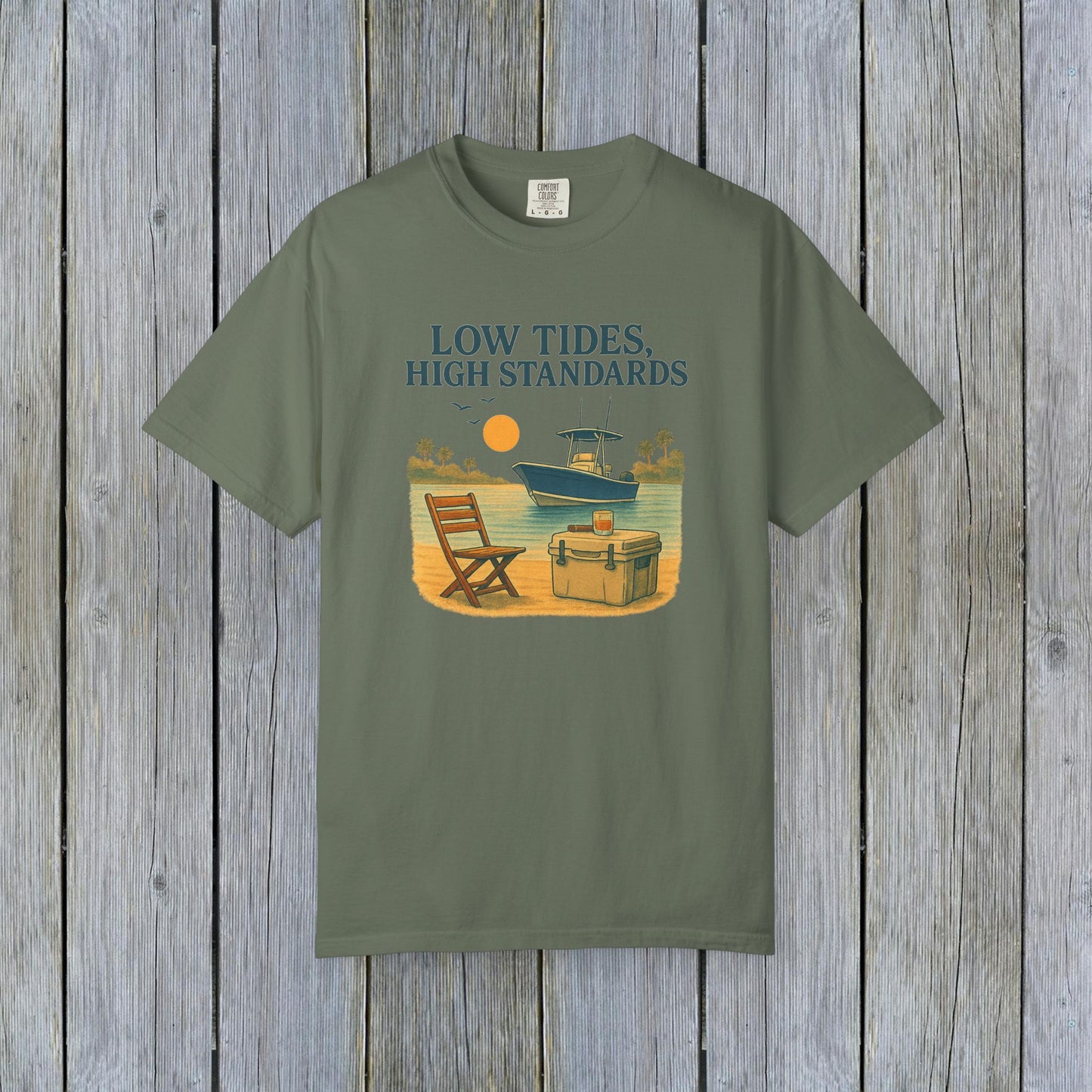 Garment-Dyed Low Tides High Standards T-Shirt in olive green color, featuring an illustration of a beach scene with a chair, cooler, and a boat in the background. Made from soft 100% US cotton, available in sizes S-4XL.