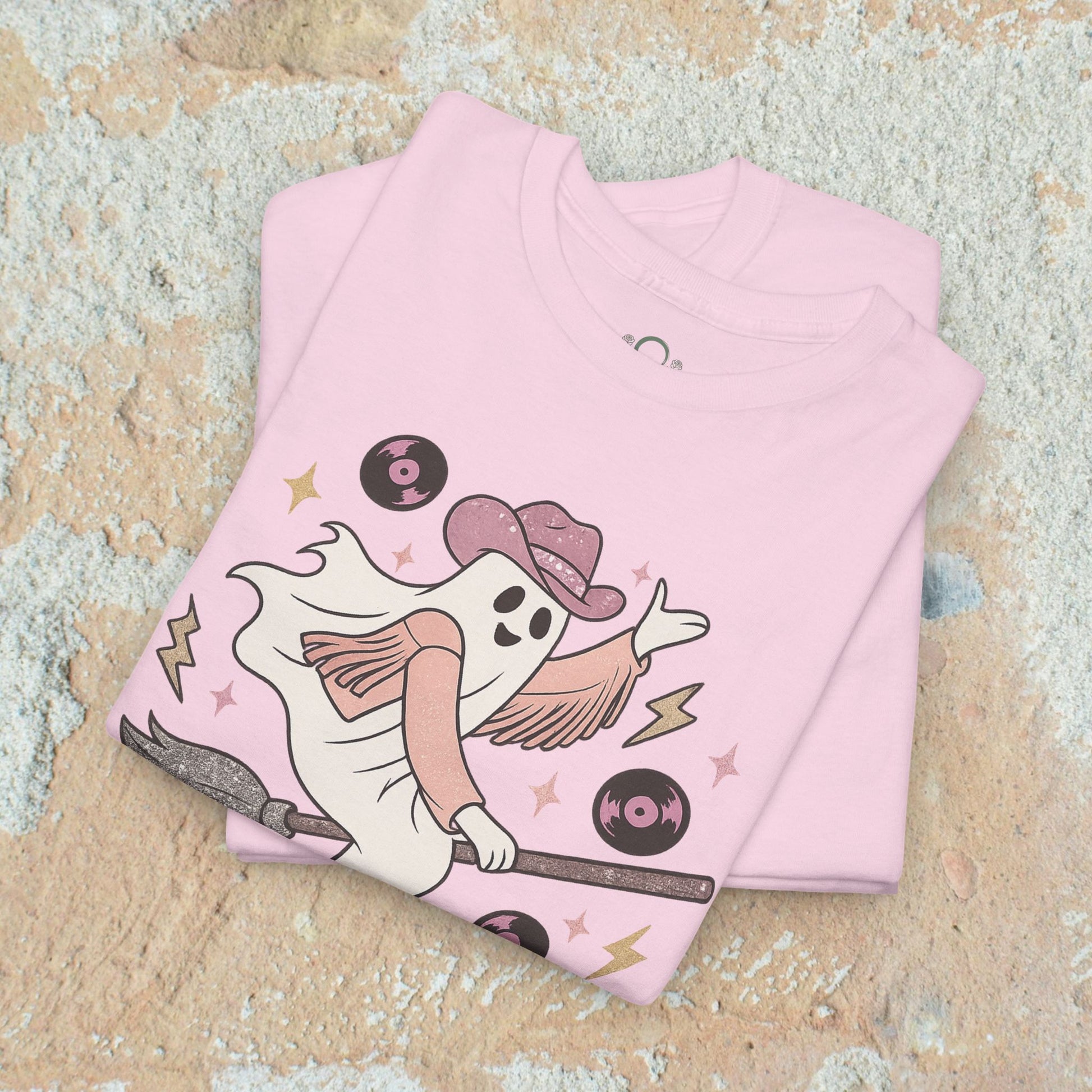 Spooky Cowgirl Graphic Tee with a fun ghost design in a stack of pink shirts, featuring a cowboy hat and broom, perfect for Halloween enthusiasts looking for soft, eco-friendly fashion.