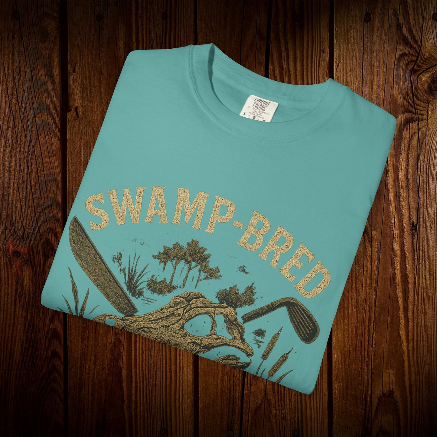 Close-up of the Unisex Swamp-Bred Fairway-Fed Tee folded, featuring a unique swamp-inspired graphic design in gold.
