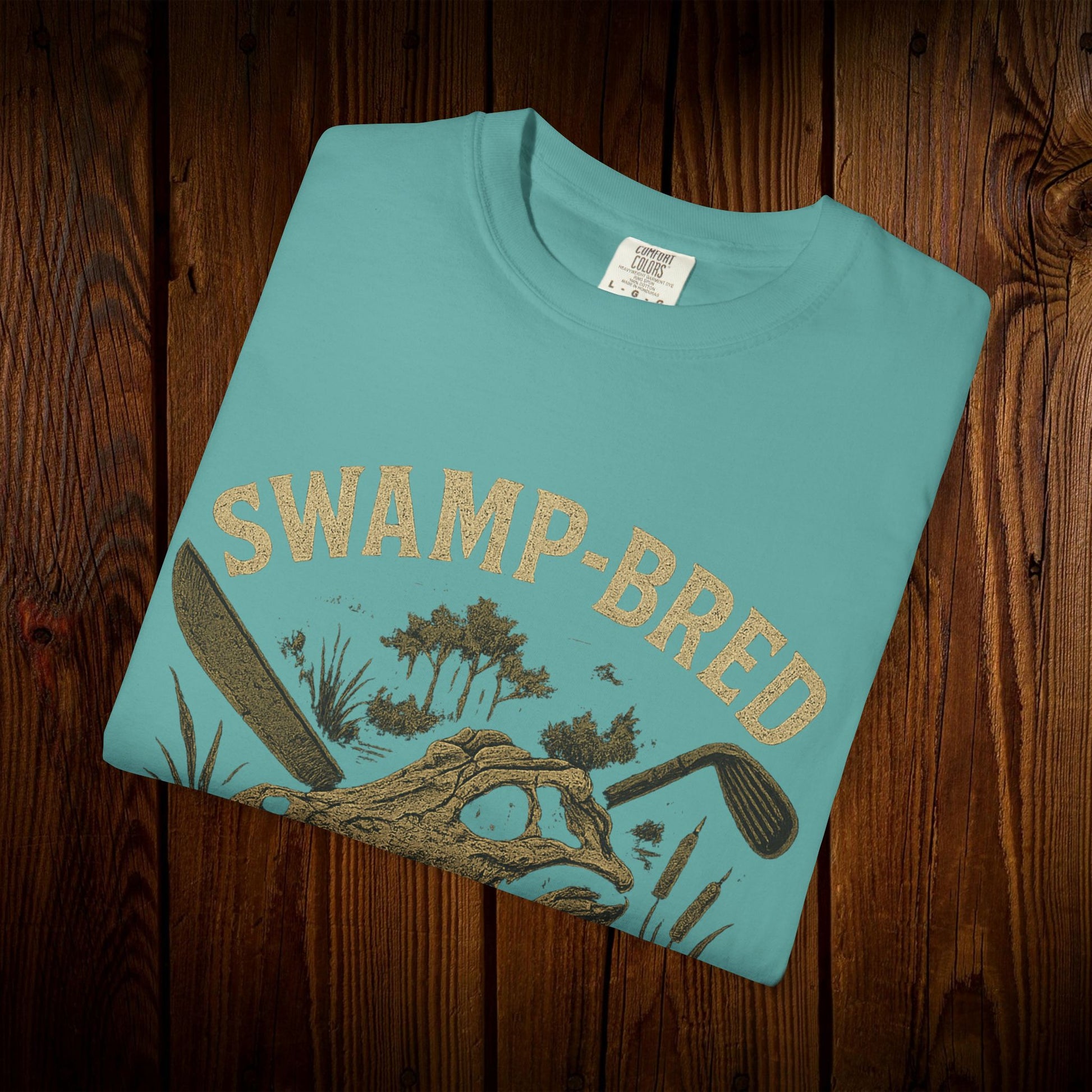 Close-up of the Unisex Swamp-Bred Fairway-Fed Tee folded, featuring a unique swamp-inspired graphic design in gold.