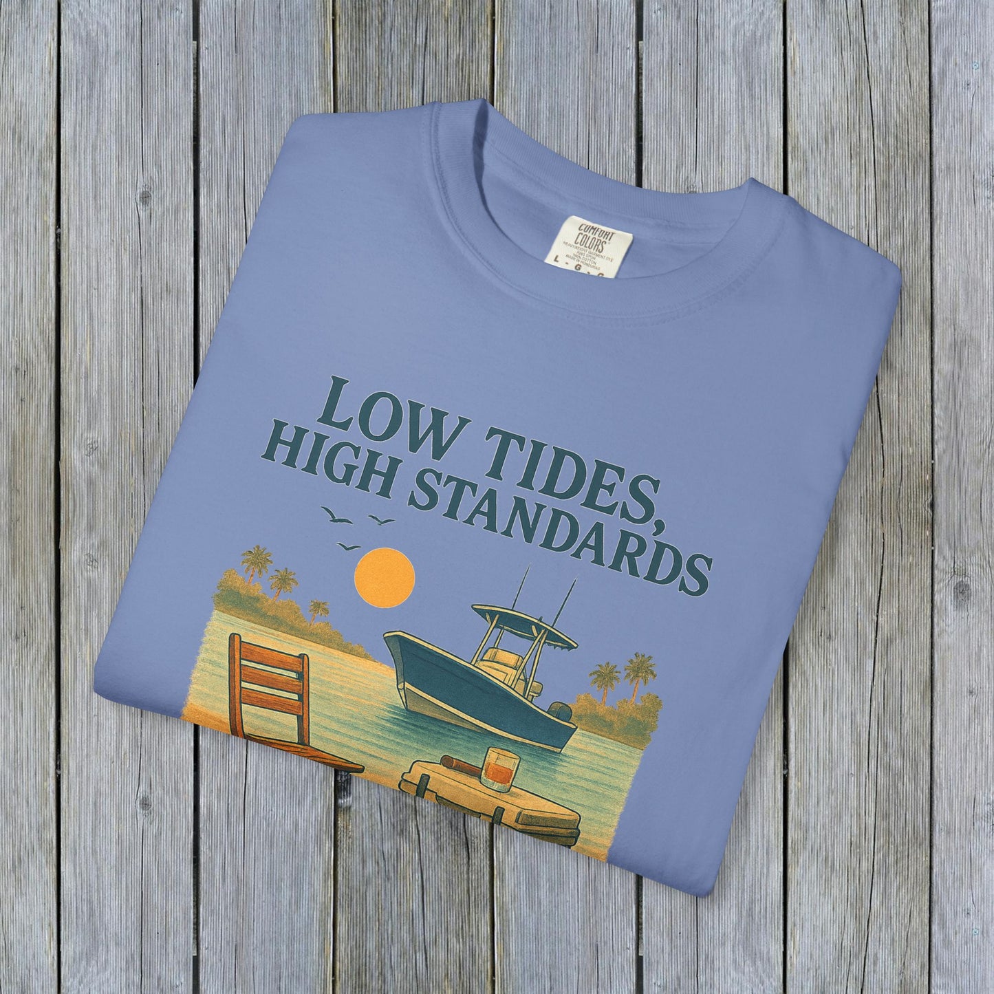 Garment-Dyed Low Tides High Standards T-Shirt displayed on wooden background, featuring a boat and sunset illustration, made of soft 100% US cotton.