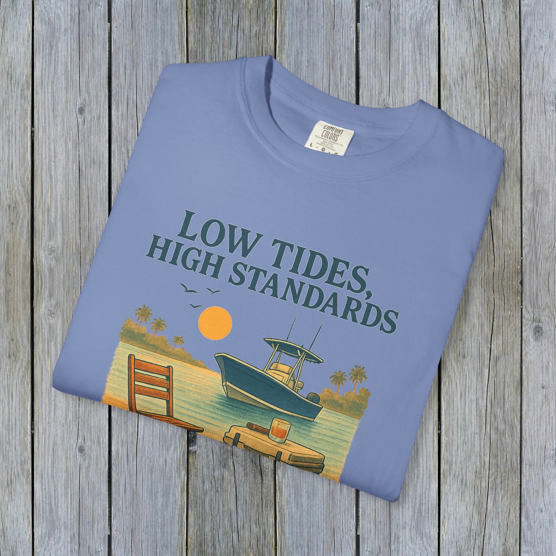 Garment-Dyed Low Tides High Standards T-Shirt displayed on wooden background, featuring a boat and sunset illustration, made of soft 100% US cotton.