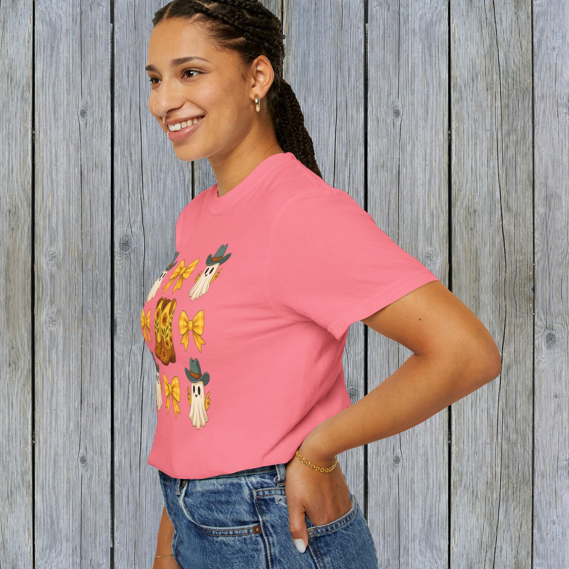 Side view of a woman wearing a Cute Halloween T-Shirt adorned with ghosts and cowboy boots, smiling against a wooden backdrop, emphasizing its charm in unisex fall fashion and eco-friendly fabric.