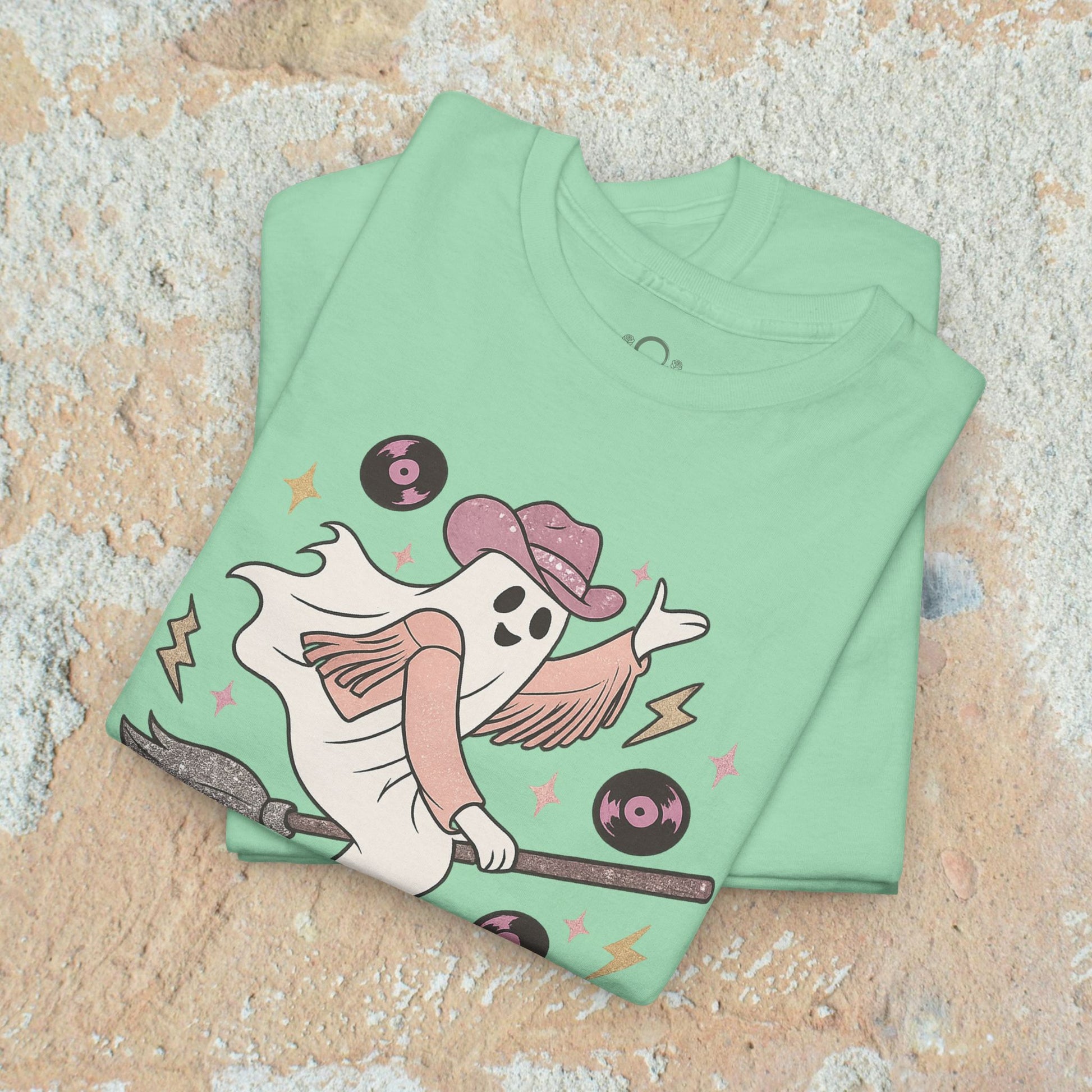 Spooky Cowgirl Graphic Tee in mint green stacked view, highlighting the fun design of a ghost cowgirl on a broomstick. Made from eco-friendly materials, sizes S-5XL available.
