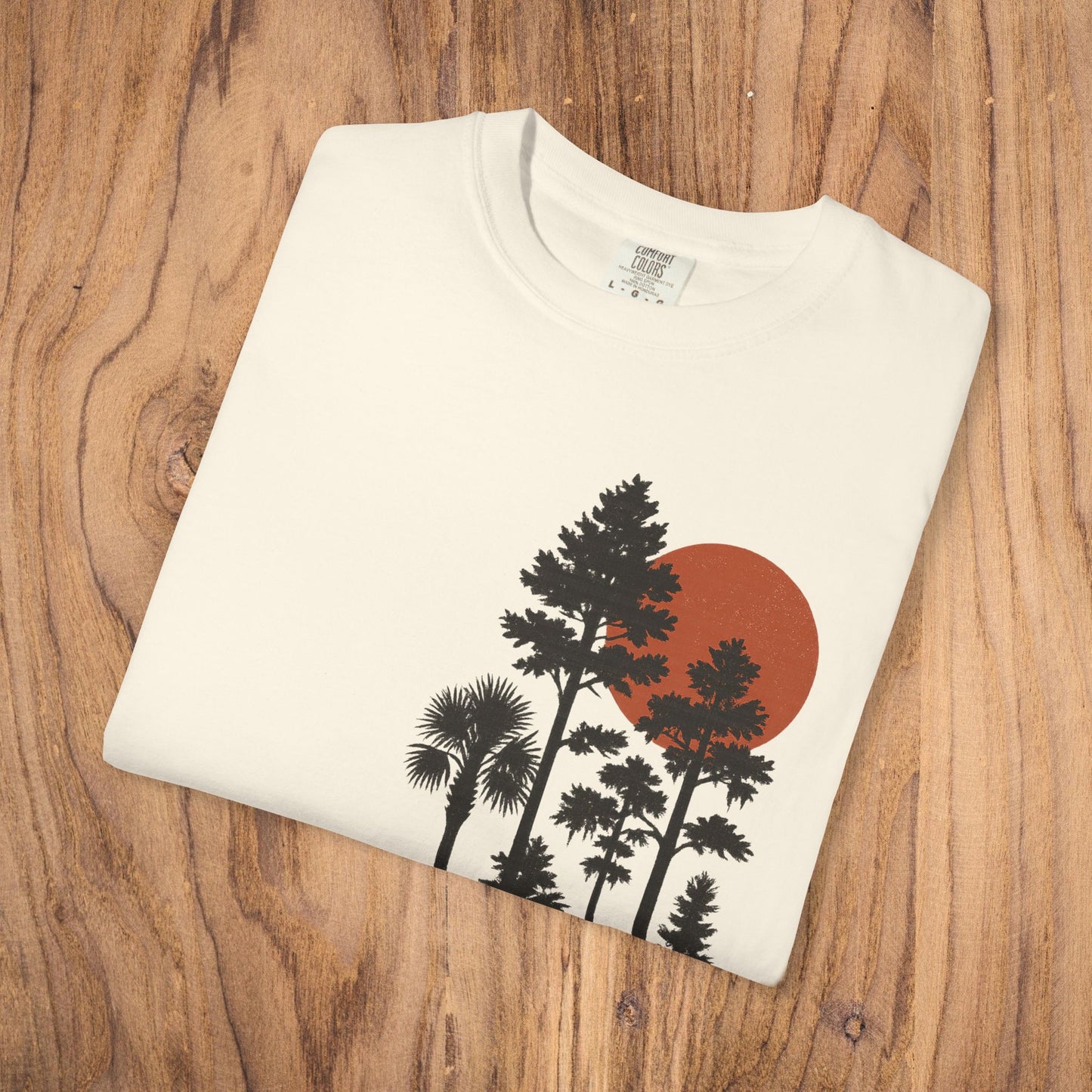 Sunset Trees Unisex T-Shirt folded on wooden surface, showcasing its soft 100% cotton fabric and vibrant sunset design, perfect for casual outings.