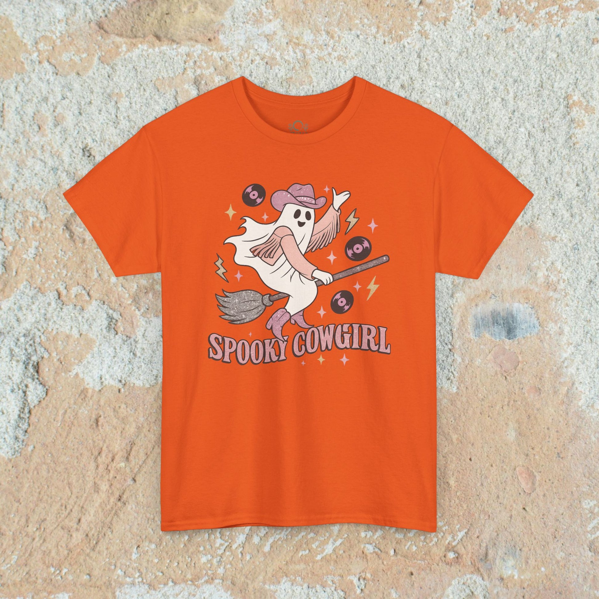 Spooky Cowgirl Graphic Tee displayed in bright orange, featuring a spirited ghost with a cowboy hat and broomstick, ideal for Halloween fashion.