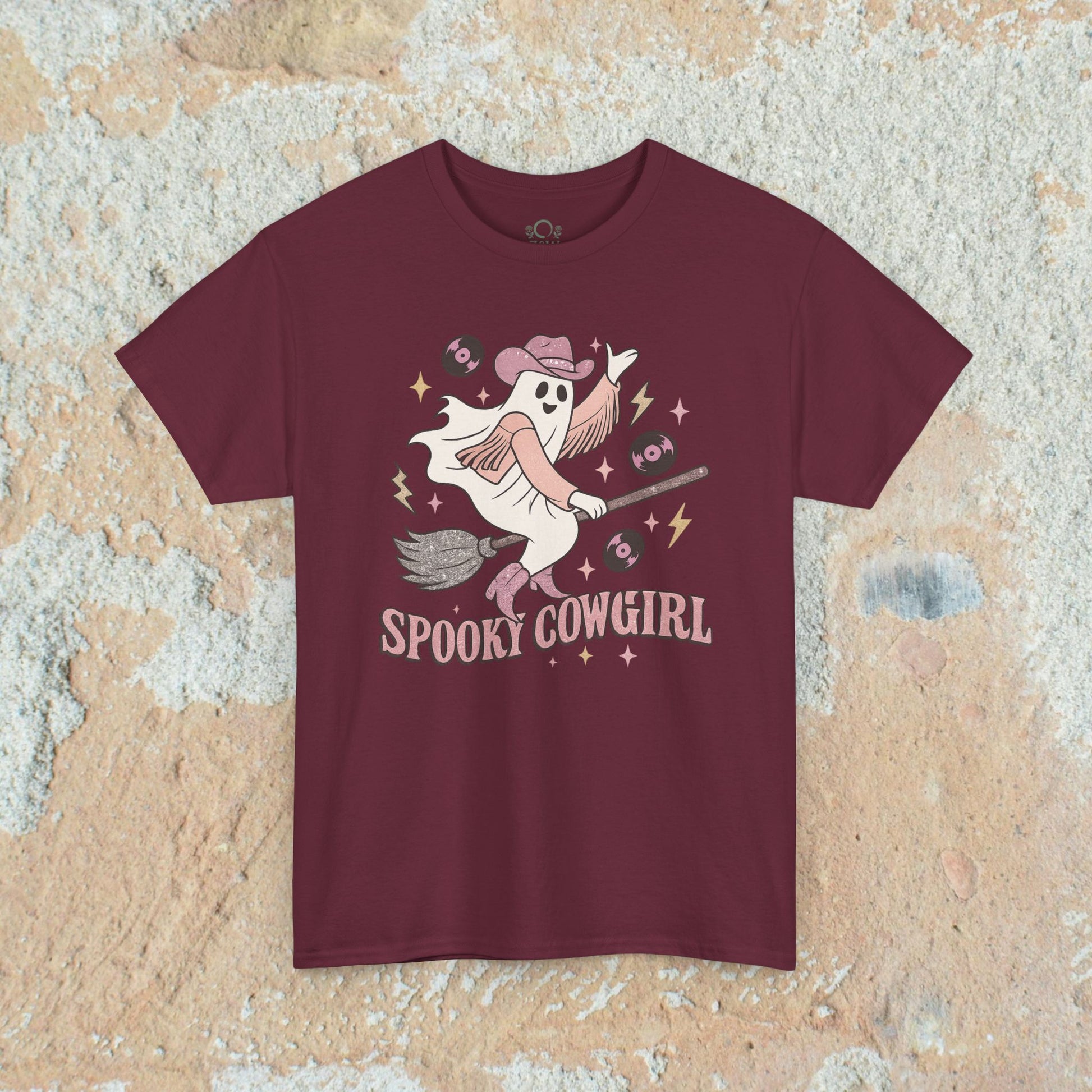 Spooky Cowgirl Graphic Tee in maroon displaying a cute ghost in a cowboy hat riding a broom, surrounded by music notes and vinyl records, made from sustainable materials.