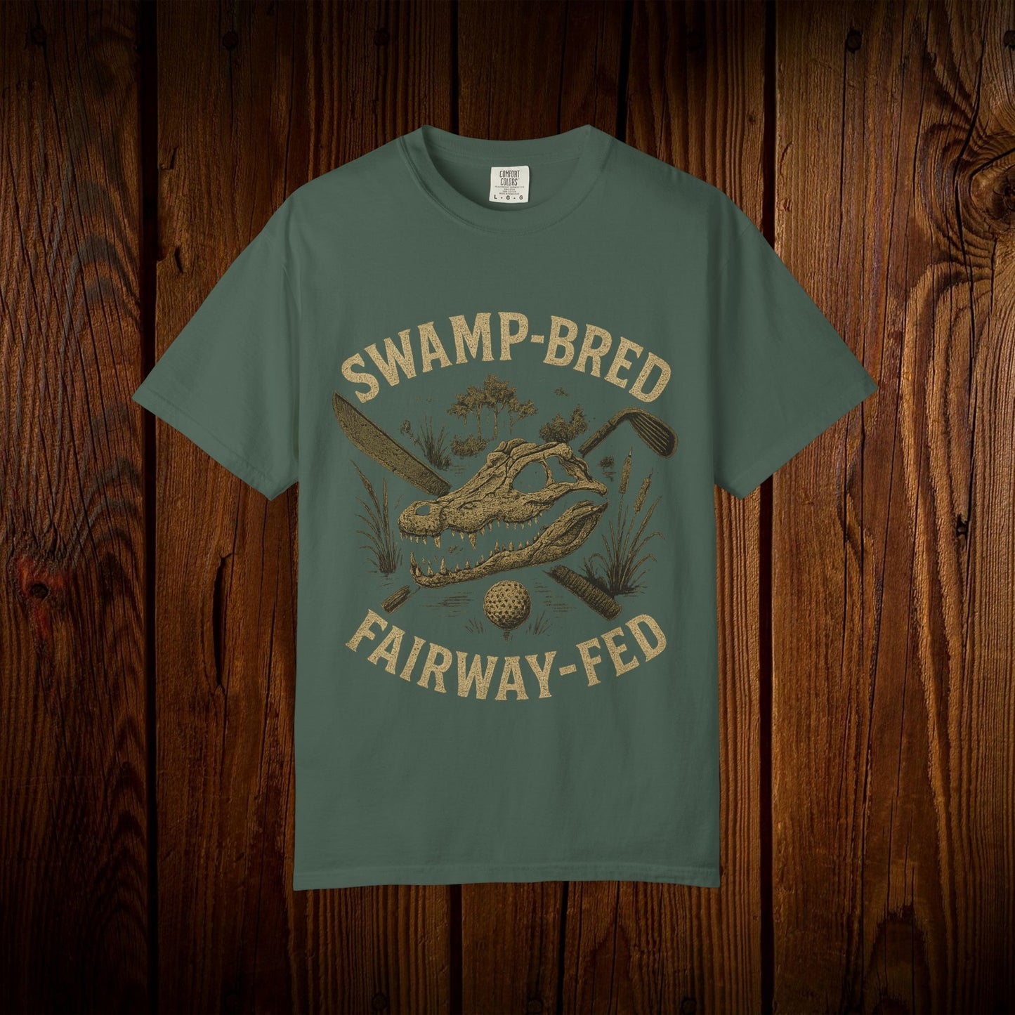 Unisex Swamp-Bred Fairway-Fed Tee in olive green, showcasing a striking illustration of an alligator skull and golf clubs against a wooden backdrop.