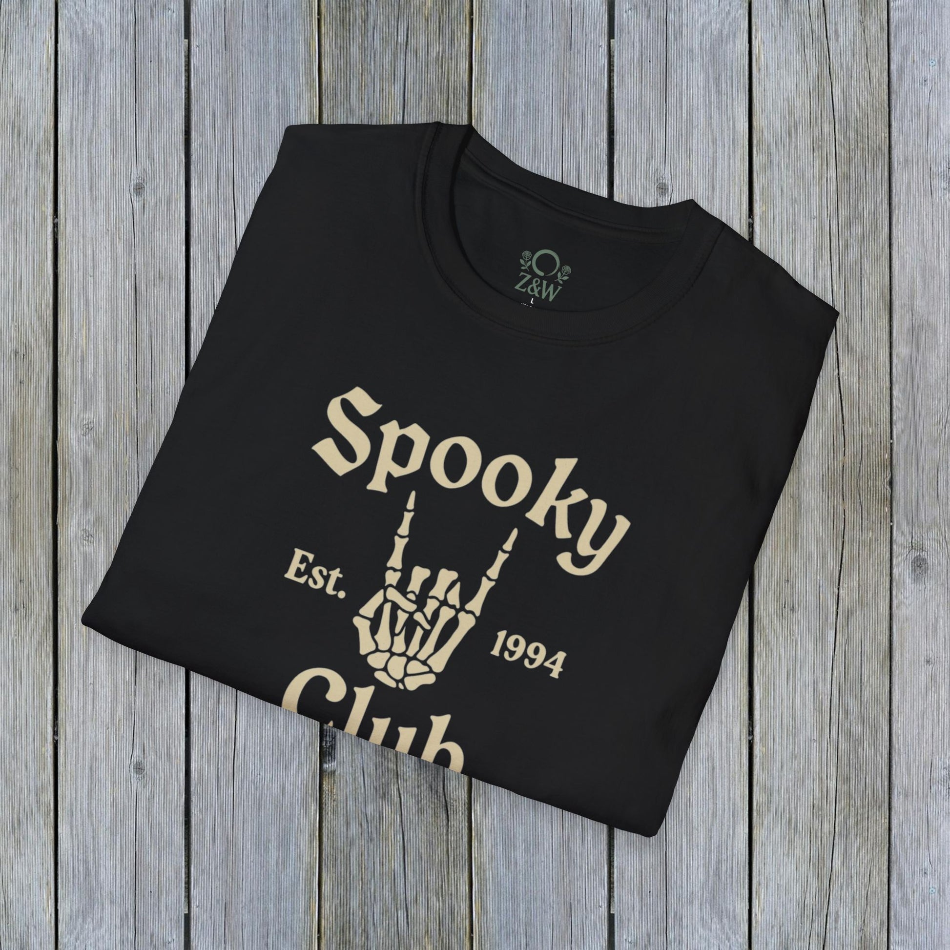 Folded Unisex Spooky Club Tee on a wooden surface, showcasing the skeleton hand design and 'Spooky Club Est. 1994' text. Made from soft, durable 100% cotton, ideal for Halloween celebrations.