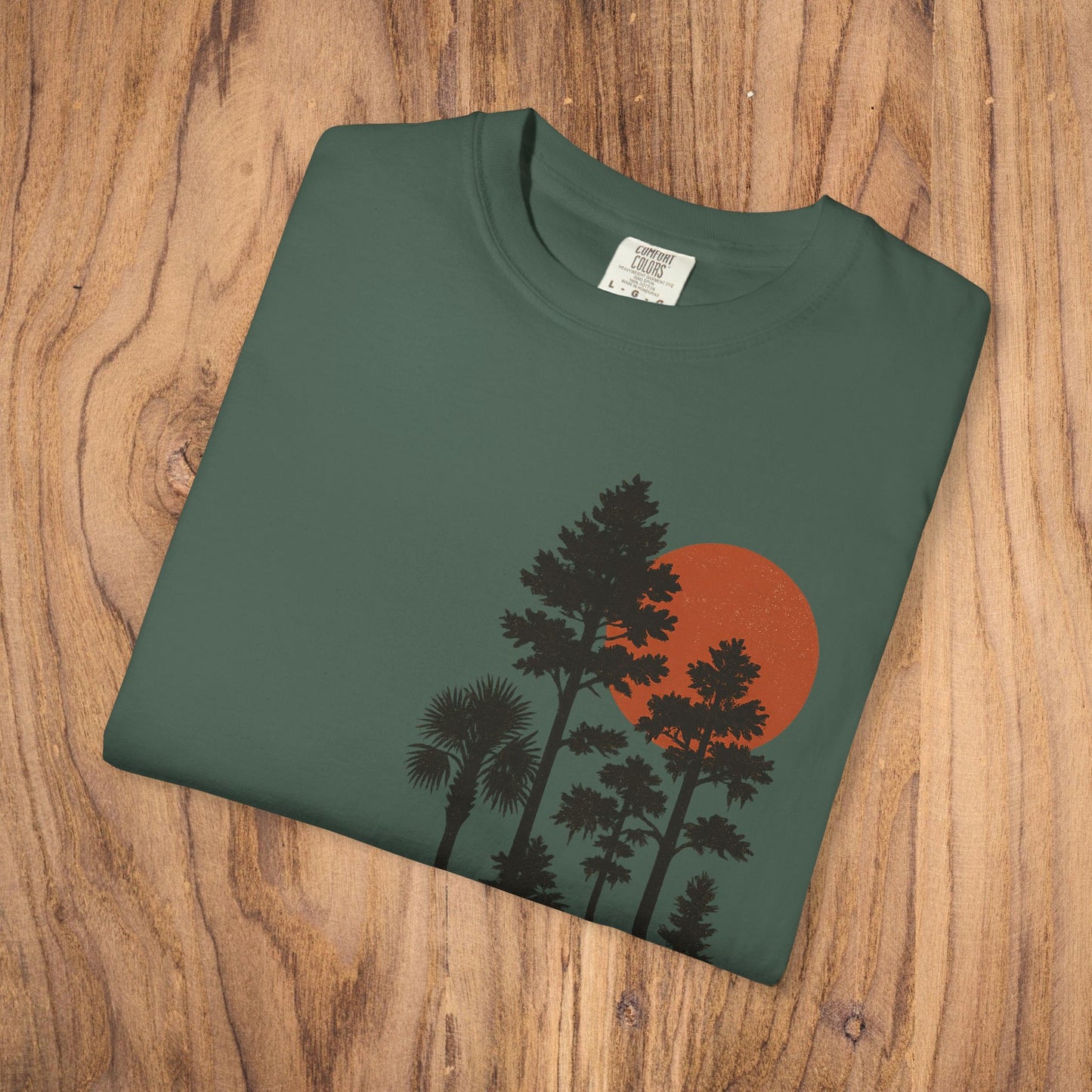 Sunset Trees Unisex T-Shirt displayed flat on wood background, showcasing the unique garment-dyed fabric and stylish design.