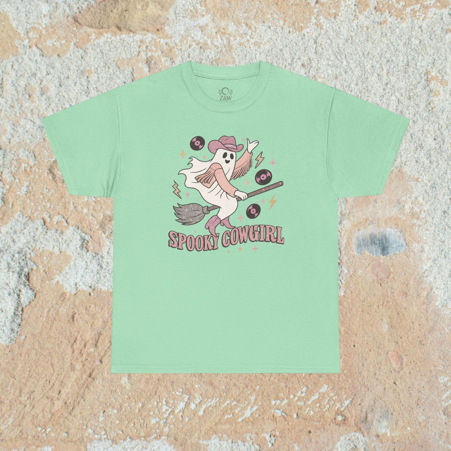Soft mint green Spooky Cowgirl graphic tee showcasing a whimsical ghost riding a broomstick, adorned with musical records, ideal for festive occasions. Sizes range from S to 5XL.