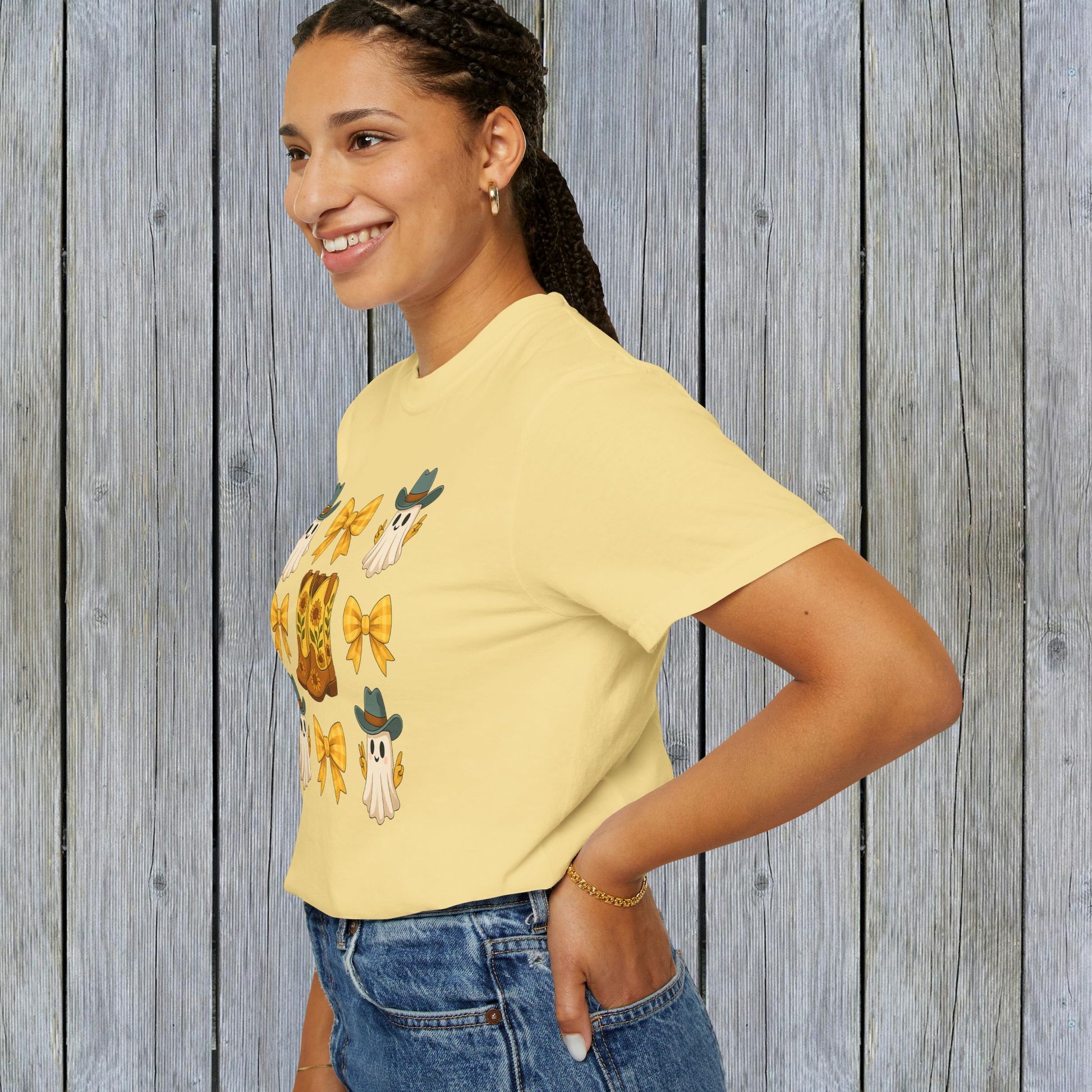 Cute Halloween T-Shirt modeled by a smiling woman, showcasing a whimsical design of ghosts and cowboy boots against a rustic wooden background, ideal for unisex fall fashion in sustainable fabric.