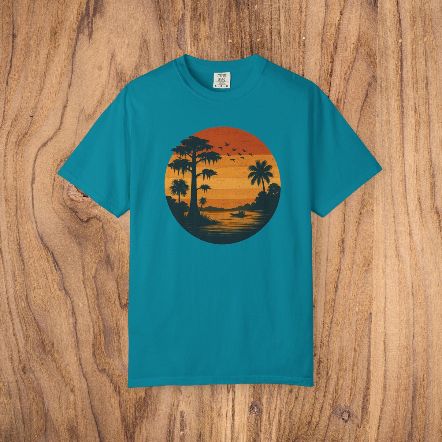 Unisex Garment-Dyed Sunset Tee in turquoise with a striking sunset illustration that includes palm trees and birds. Made from soft 100% cotton, available in sizes S to 4XL.