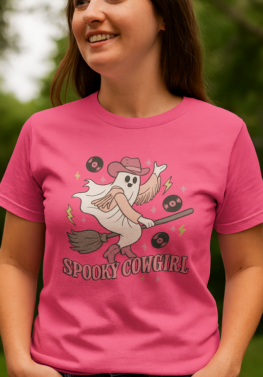 A woman wearing a Spooky Cowgirl Graphic Tee, featuring a playful ghost character dressed as a cowgirl, complete with a hat and broomstick, set against a vibrant pink background. This eco-friendly tee comes in sizes S-5XL, crafted from soft fabric for comfort.