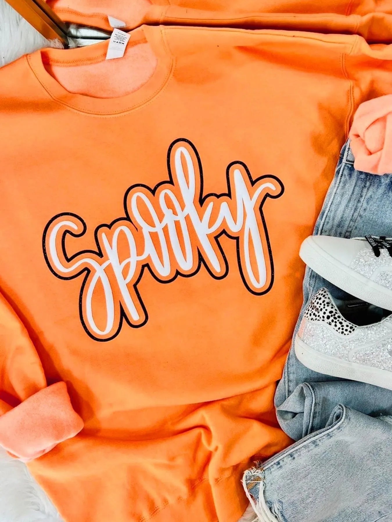 Flat lay of the Chic Spooky Round Neck Long Sleeve Sweatshirt in orange, paired with denim jeans and trendy sneakers, perfect for Halloween.