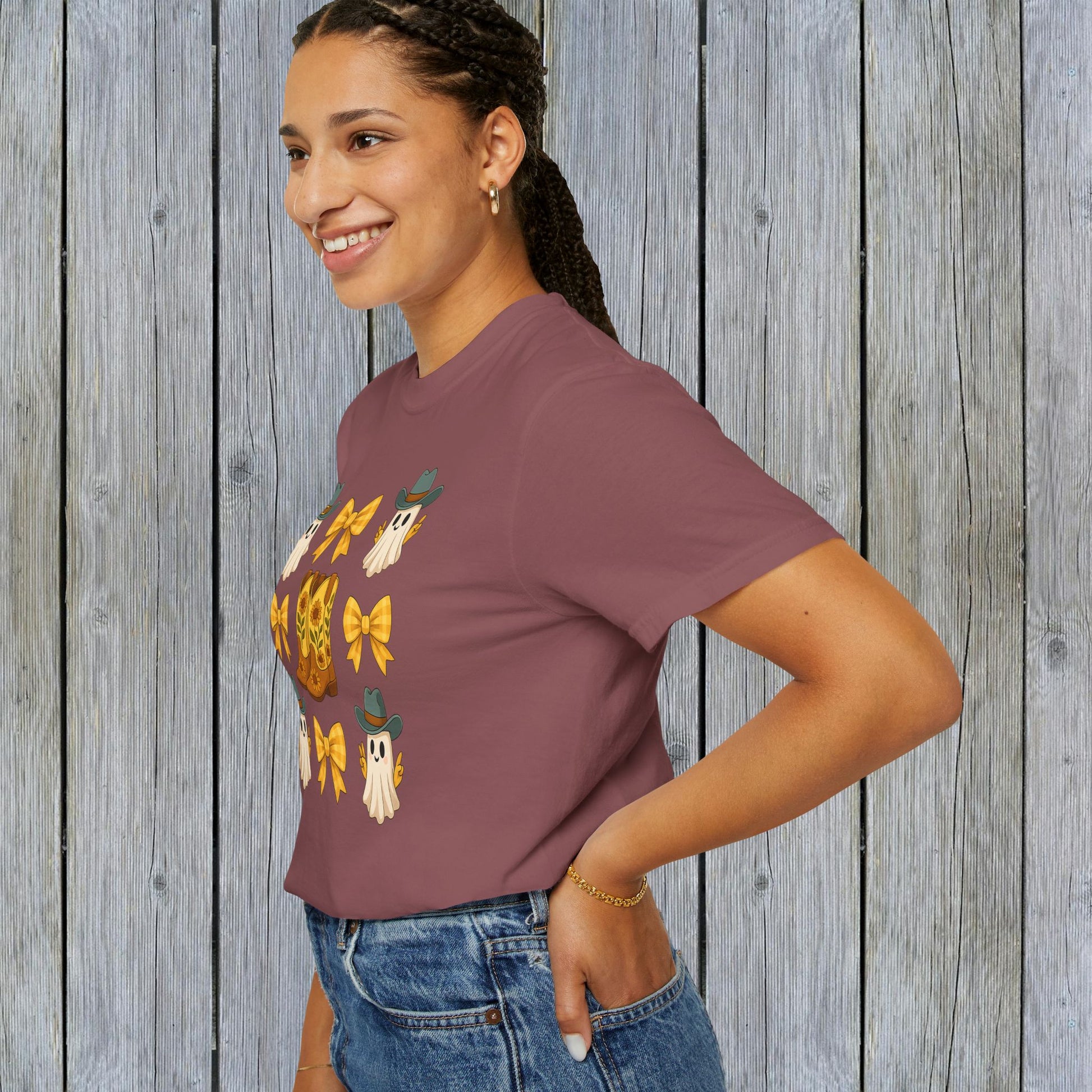 Cute Halloween T-Shirt designed with ghosts and cowboy boots, worn by a woman in maroon against a rustic wooden backdrop. A trendy option for unisex fall fashion made from eco-friendly fabric.