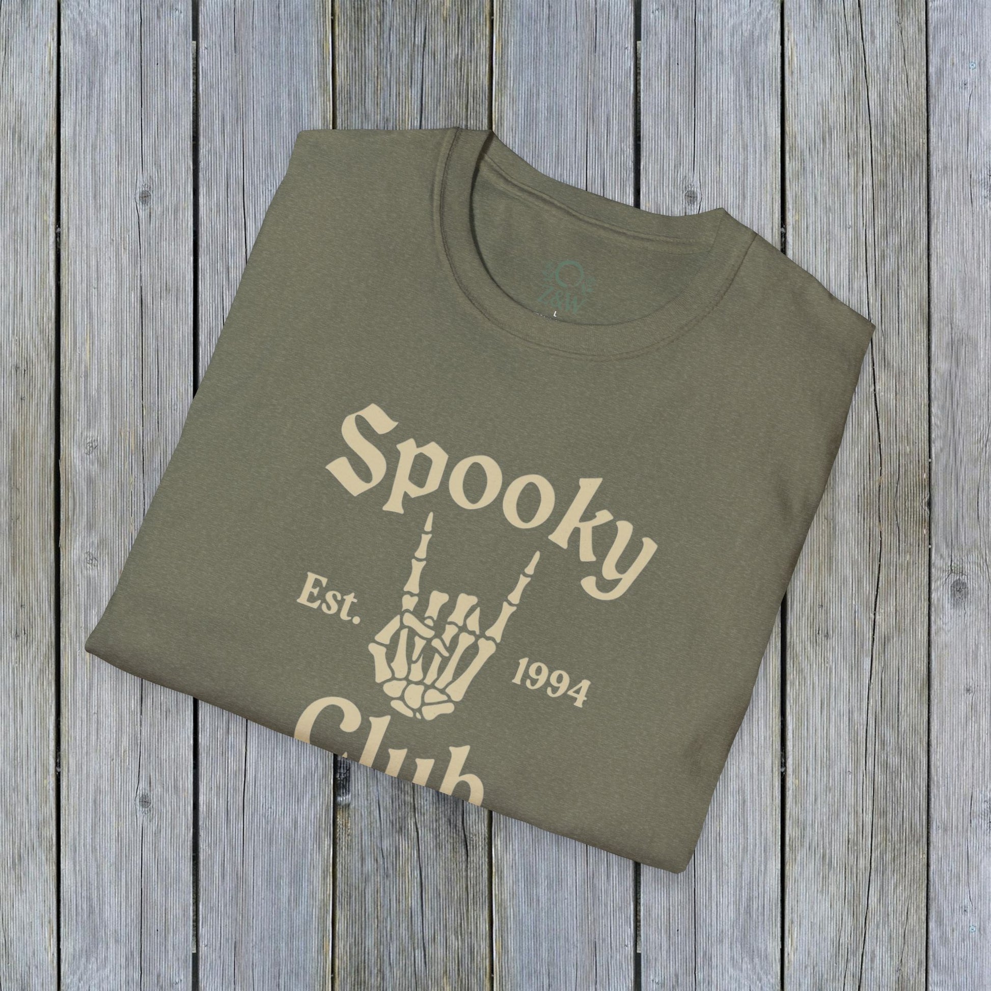 Folded Unisex Spooky Club Tee in olive green, set against a wooden background, highlighting the unique skeleton hand graphic and 'Spooky Club Est. 1994' print. Crafted from soft, durable 100% cotton, great for Halloween outings.
