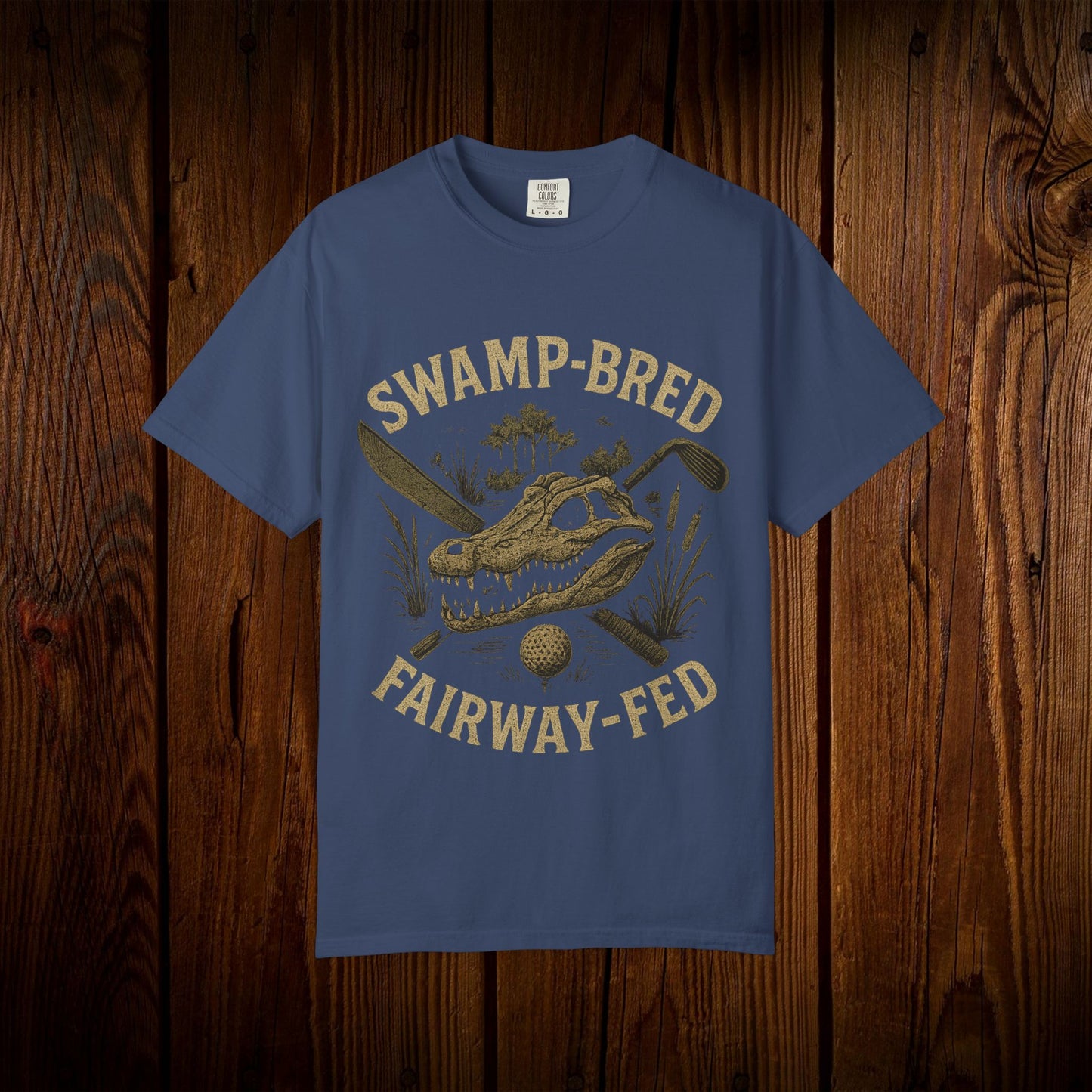 Unisex Swamp-Bred Fairway-Fed Tee in navy blue, featuring a unique alligator skull design with golf clubs, set against a warm wooden texture.