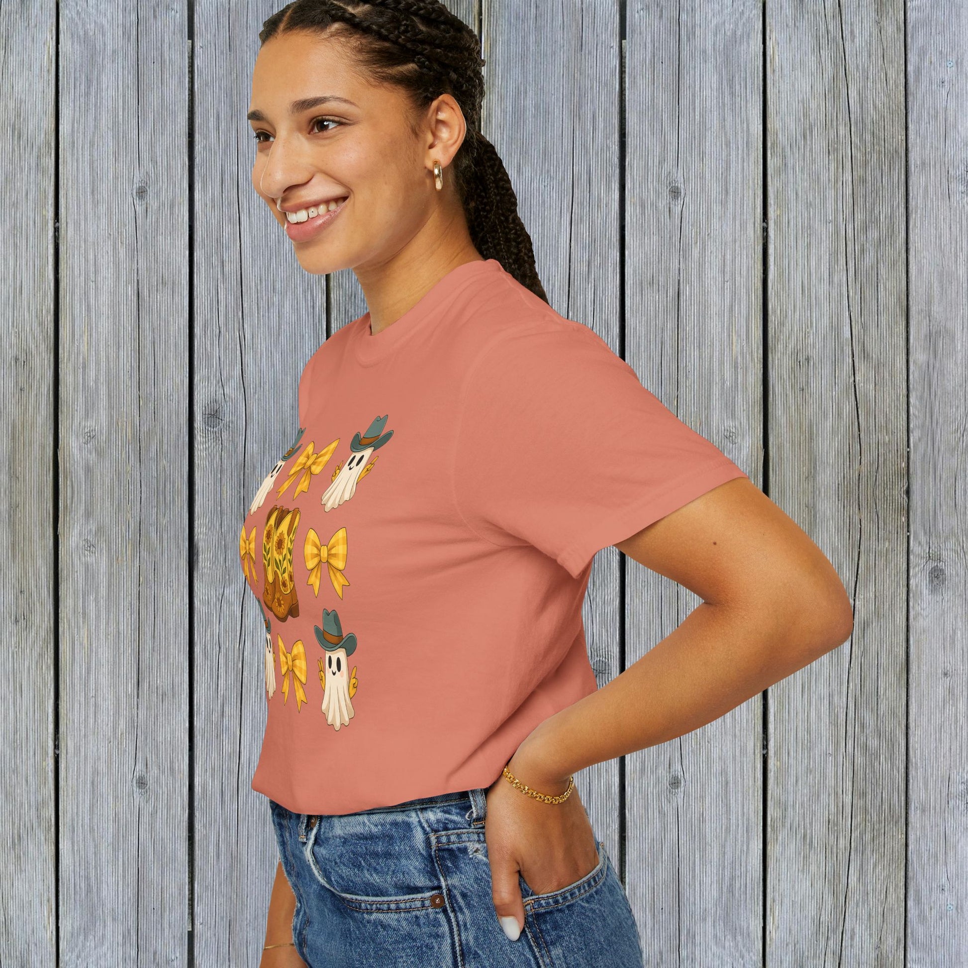 Cute Halloween T-Shirt featuring ghosts and cowboy boots, shown on a smiling woman against a rustic wooden background, perfect for unisex fall fashion in eco-friendly fabric.