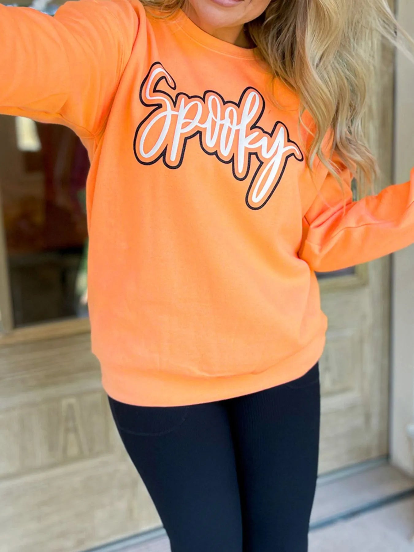 Model wearing Chic Spooky Round Neck Long Sleeve Sweatshirt in orange with 'Spooky' graphic, complemented by fitted black leggings.