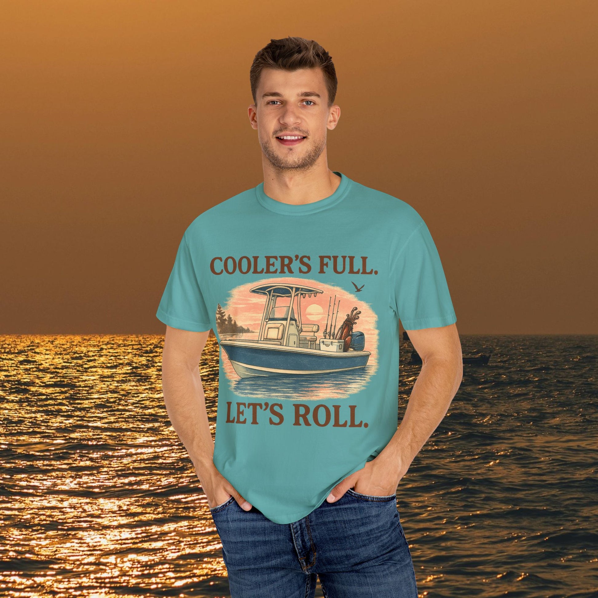 A man smiling in a turquoise Men's & Women's Durable Cotton Full Tee displaying the phrase 'COOLER'S FULL. LET'S ROLL.' while enjoying a sunset by the water.