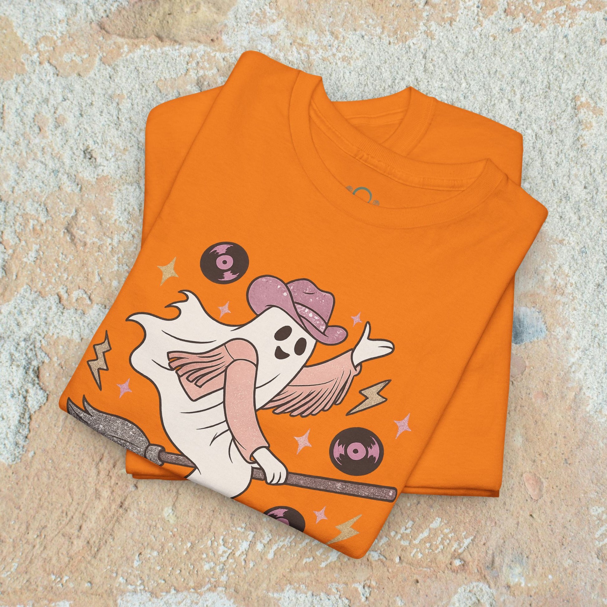 Spooky Cowgirl Graphic Tee in vibrant orange, featuring a playful ghost with a cowboy hat riding a broom. Eco-friendly and soft fabric, available in sizes S-5XL.