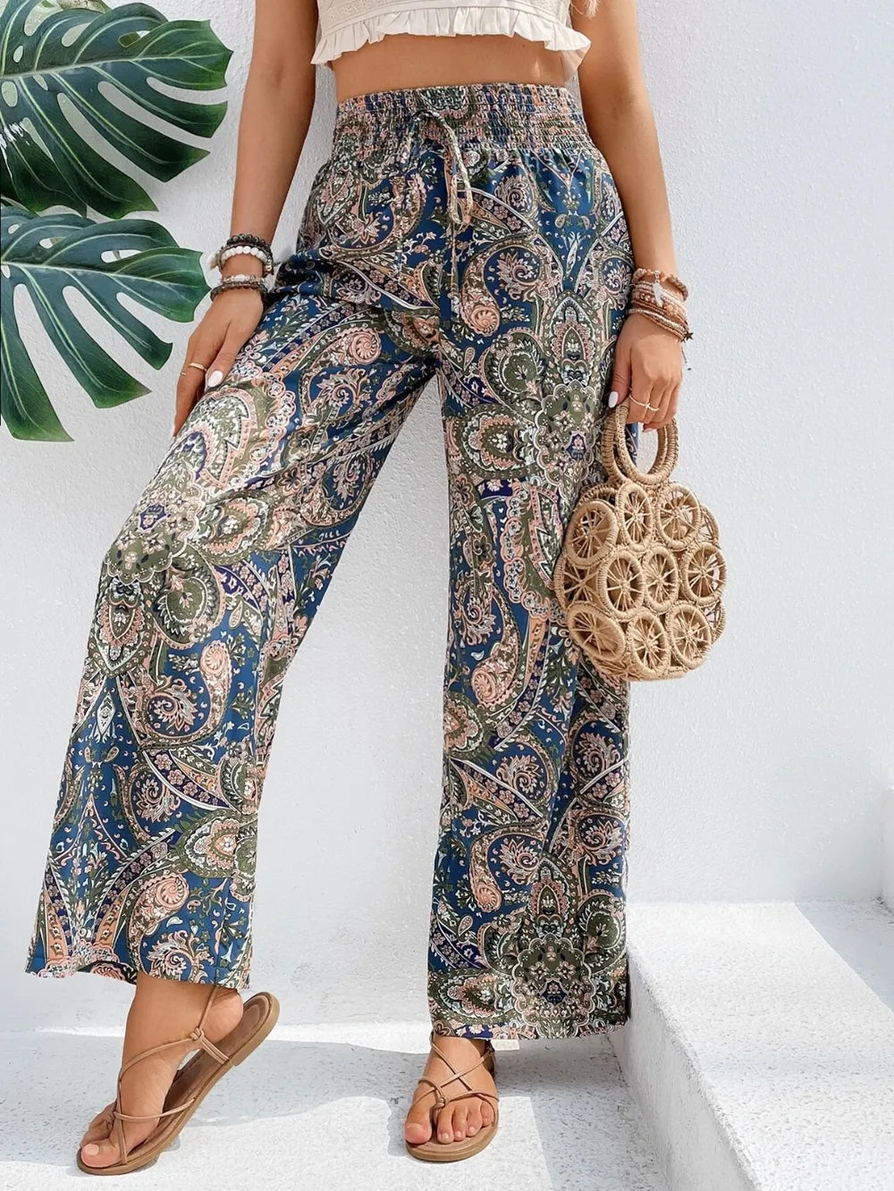 Model wears Trendy Printed Wide Leg Pants with Tied Detail, beautifully paired with a woven bag, highlighting the elegant paisley print made from 100% opaque polyester.