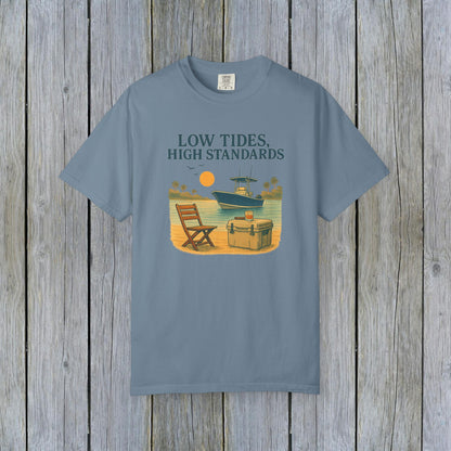 Garment-Dyed Low Tides High Standards T-Shirt in blue color, showcasing a vibrant beach illustration with a chair and cooler beside a boat. This soft 100% US cotton tee is offered in sizes S-4XL.