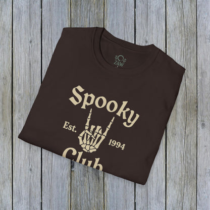 Folded Unisex Spooky Club Tee in dark brown, placed on a wooden backdrop, featuring spooky skeleton hand artwork and 'Spooky Club Est. 1994' tagline. Soft 100% cotton fabric perfect for Halloween.