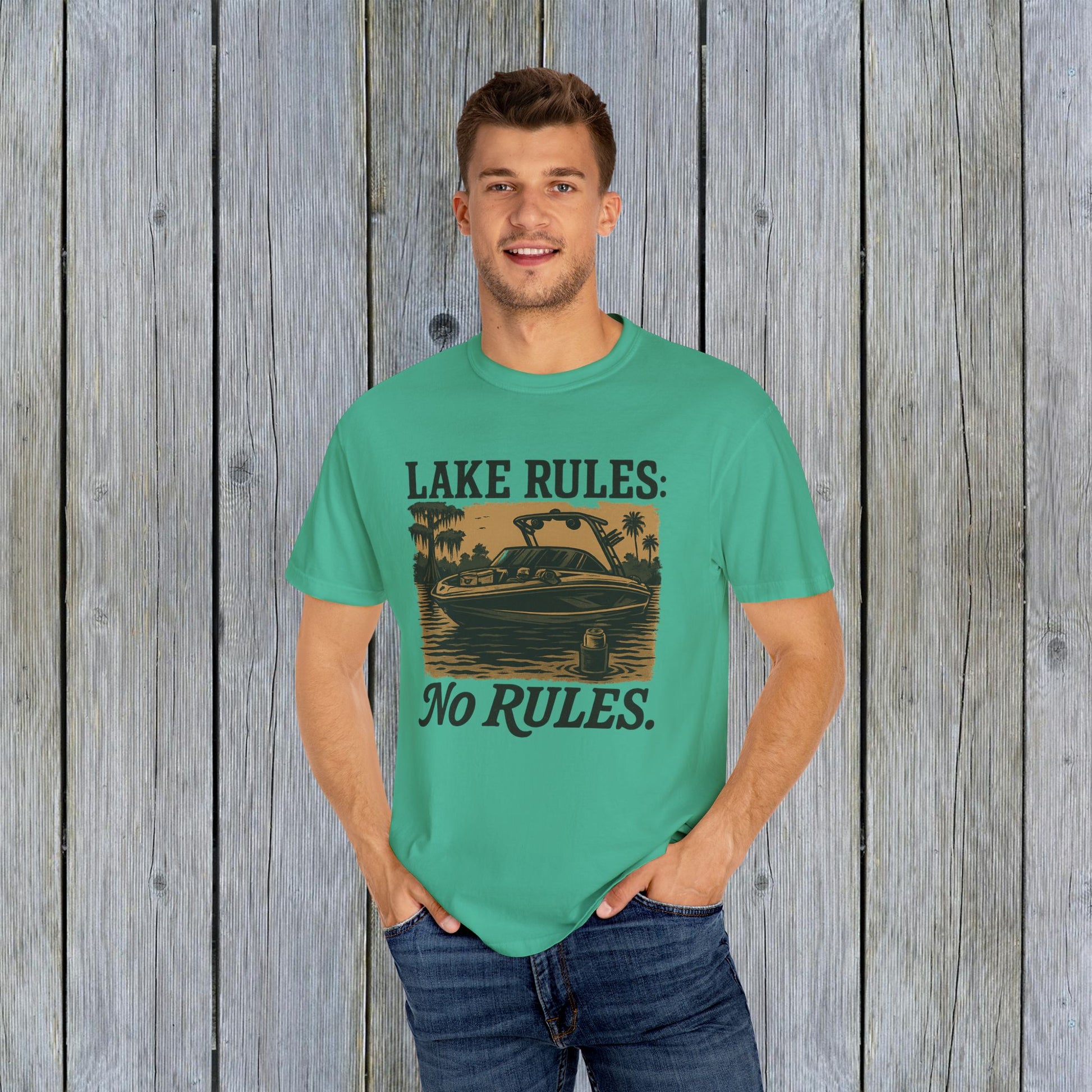 Young man wearing the Unisex Garment-Dyed Lake Rules Tee in bright teal, showcasing its relaxed fit and 'No Rules' boat graphic against a rustic wooden backdrop.