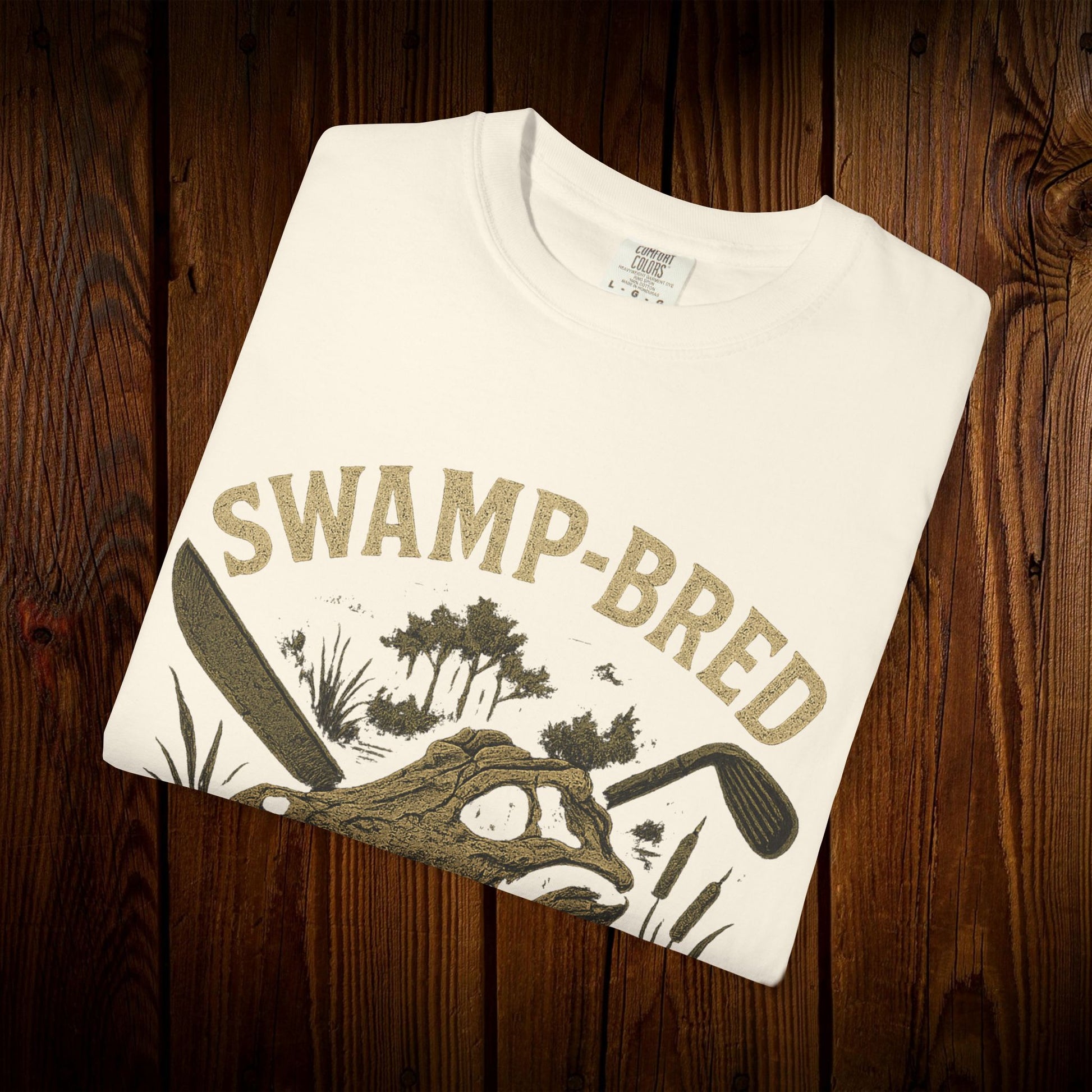 Close-up of the Unisex Swamp-Bred Fairway-Fed Tee folded in white, featuring the eye-catching swamp-themed graphic design in gold.