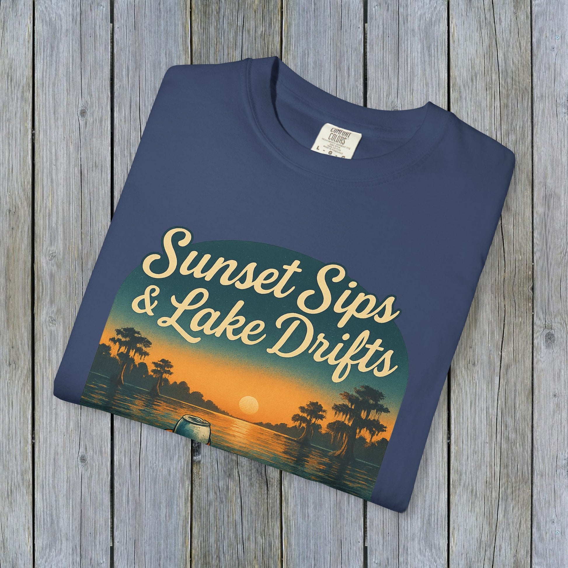 Folded Lake Drifts T-Shirt in deep navy color, showcasing the 'Sunset Sips & Lake Drifts' artwork on a wooden surface.