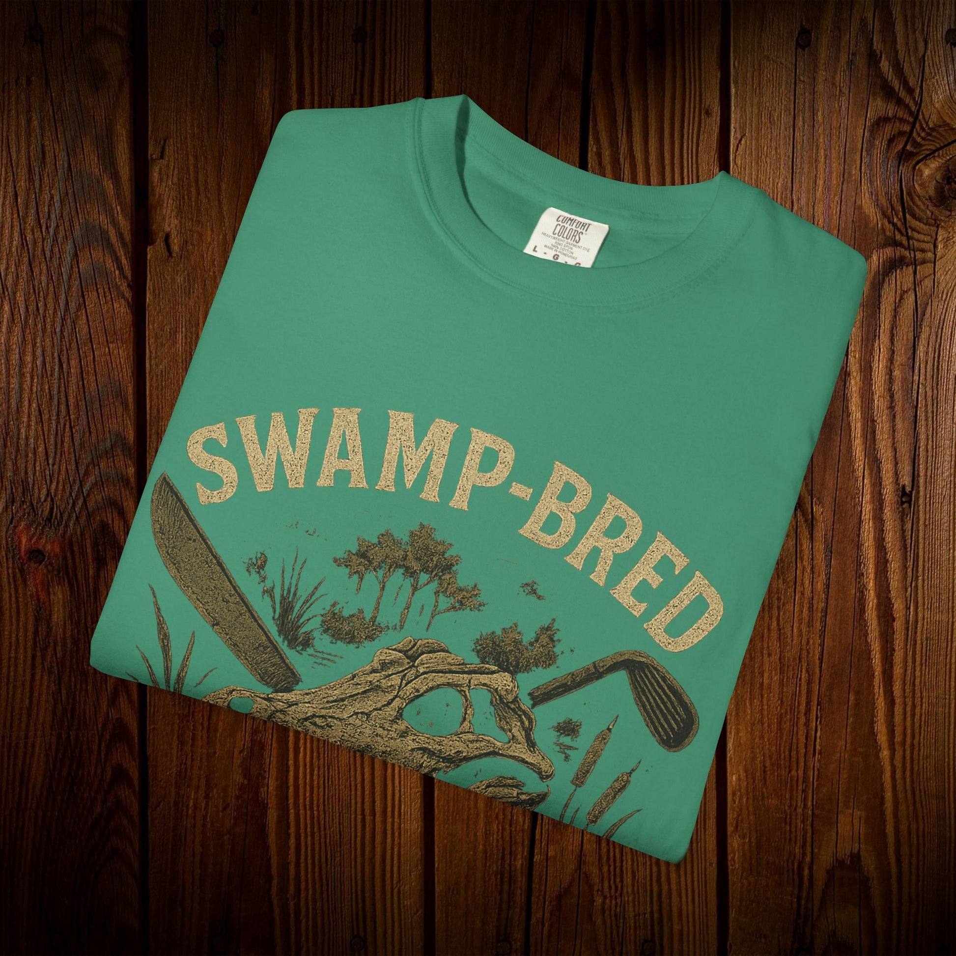 Folded Unisex Swamp-Bred Fairway-Fed Tee in a striking teal color, laid out on a wooden surface. Made from 100% pre-shrunk US cotton with detailed graphics that appeal to golf fans.