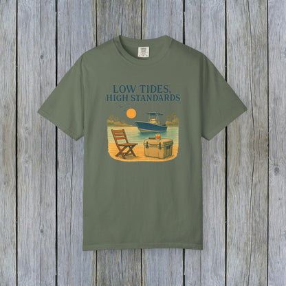 Garment-Dyed Low Tides High Standards T-Shirt in olive green color, featuring an illustration of a beach scene with a chair, cooler, and a boat in the background. Made from soft 100% US cotton, available in sizes S-4XL.