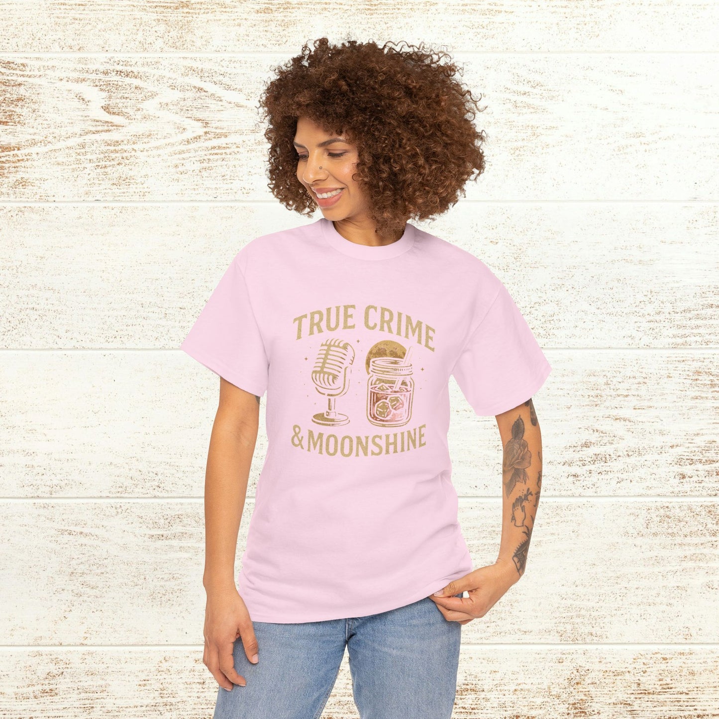 Model wearing True Crime & Moonshine Unisex Graphic Tee in soft pink against a rustic wooden backdrop, highlighting its stylish and casual vibe for true crime enthusiasts.