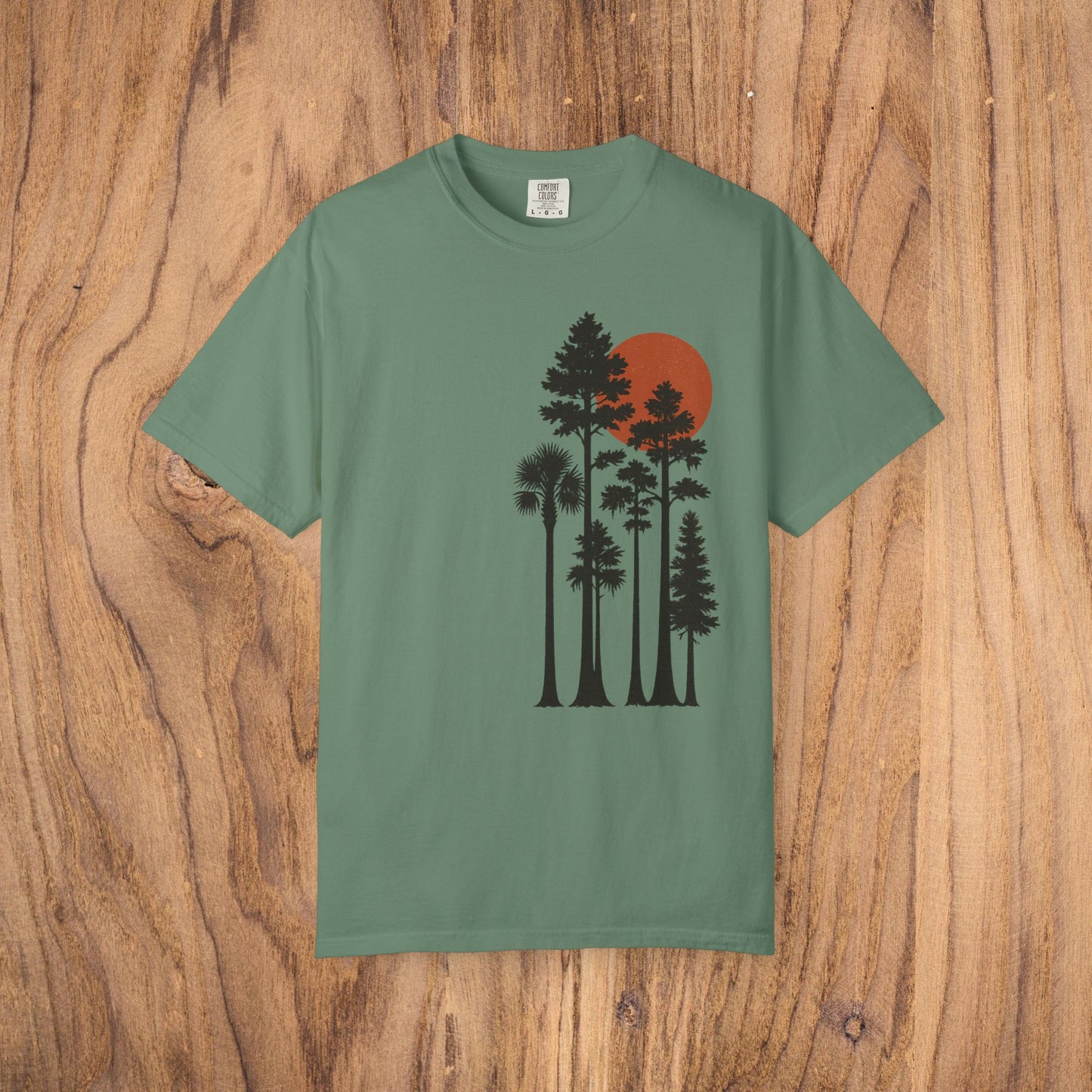 Sunset Trees Unisex T-Shirt in dusty green, constructed from soft 100% cotton in a garment-dyed style, resting on a wooden surface.