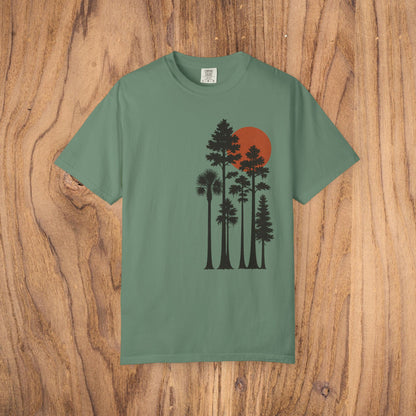 Sunset Trees Unisex T-Shirt in dusty green, constructed from soft 100% cotton in a garment-dyed style, resting on a wooden surface.