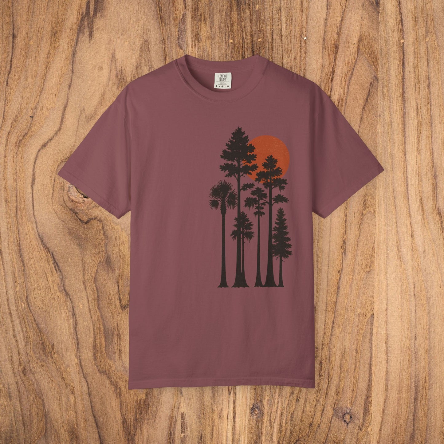 Sunset Trees Unisex T-Shirt in maroon, crafted from high-quality 100% cotton with a stylish garment-dyed texture, displayed on a natural wood background.