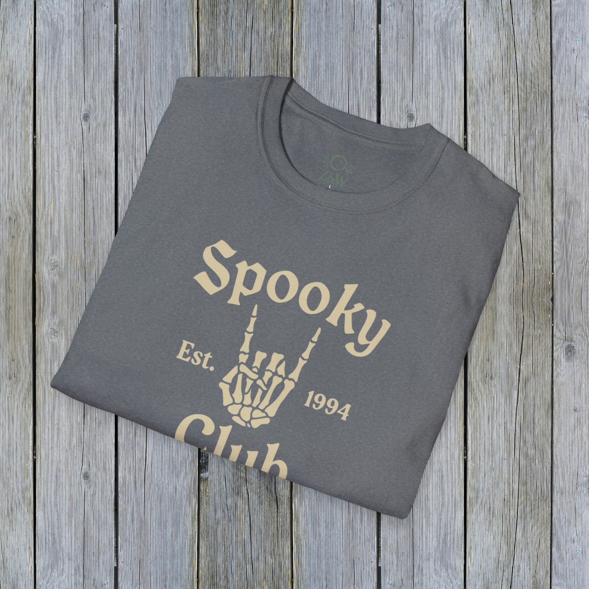 Folded Unisex Spooky Club Tee in heather gray, resting on a rustic wooden surface. The tee features a skeleton hand and 'Spooky Club Est. 1994' text, made from soft 100% cotton for a comfy Halloween wear.