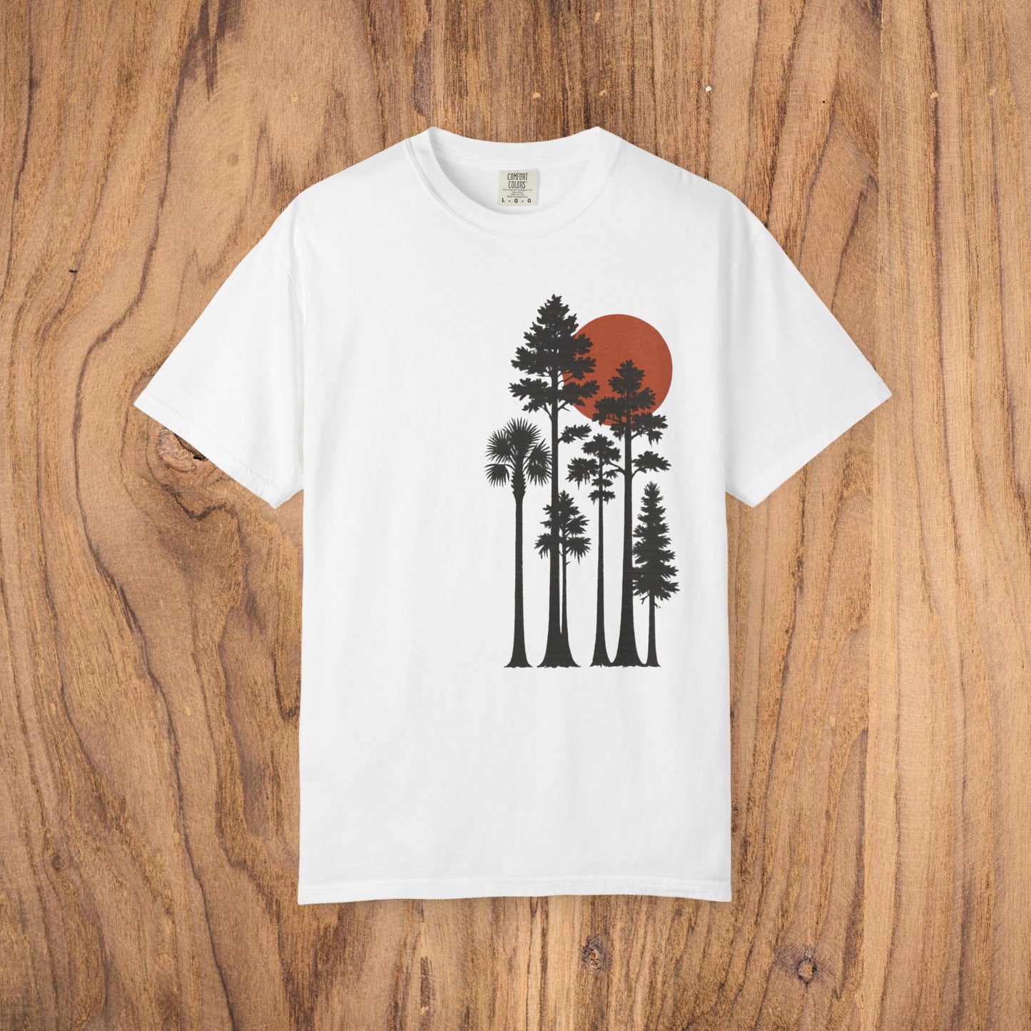 Sunset Trees Unisex T-Shirt in classic white, made from soft 100% cotton with a unique garment-dyed finish, presented against a natural wooden backdrop.