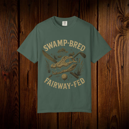Unisex Swamp-Bred Fairway-Fed Tee in olive green, showcasing a striking illustration of an alligator skull and golf clubs against a wooden backdrop.
