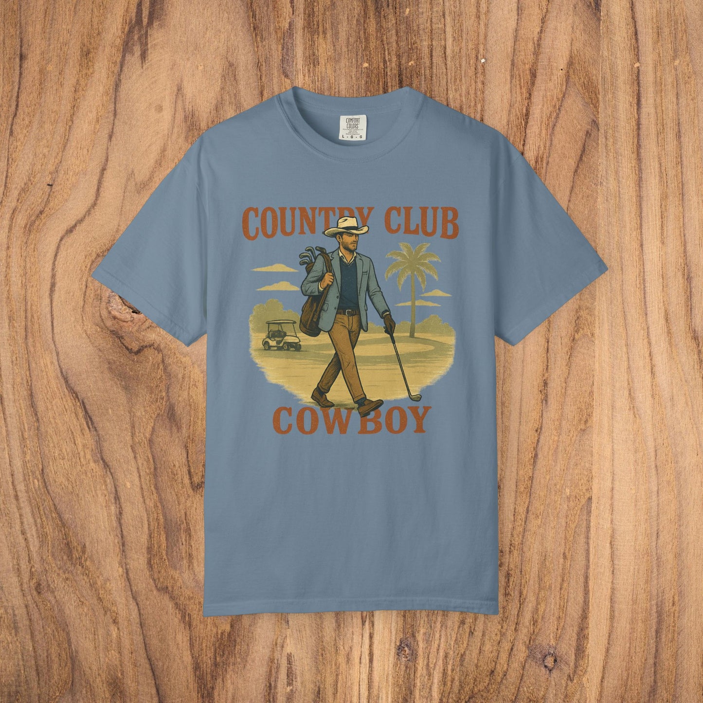 Country Club Cowboy Graphic Tee in slate blue featuring a cowboy with a golf bag set in a scenic palm tree landscape, constructed from durable cotton, equipped with ribbed collar and eco-conscious crewneck.