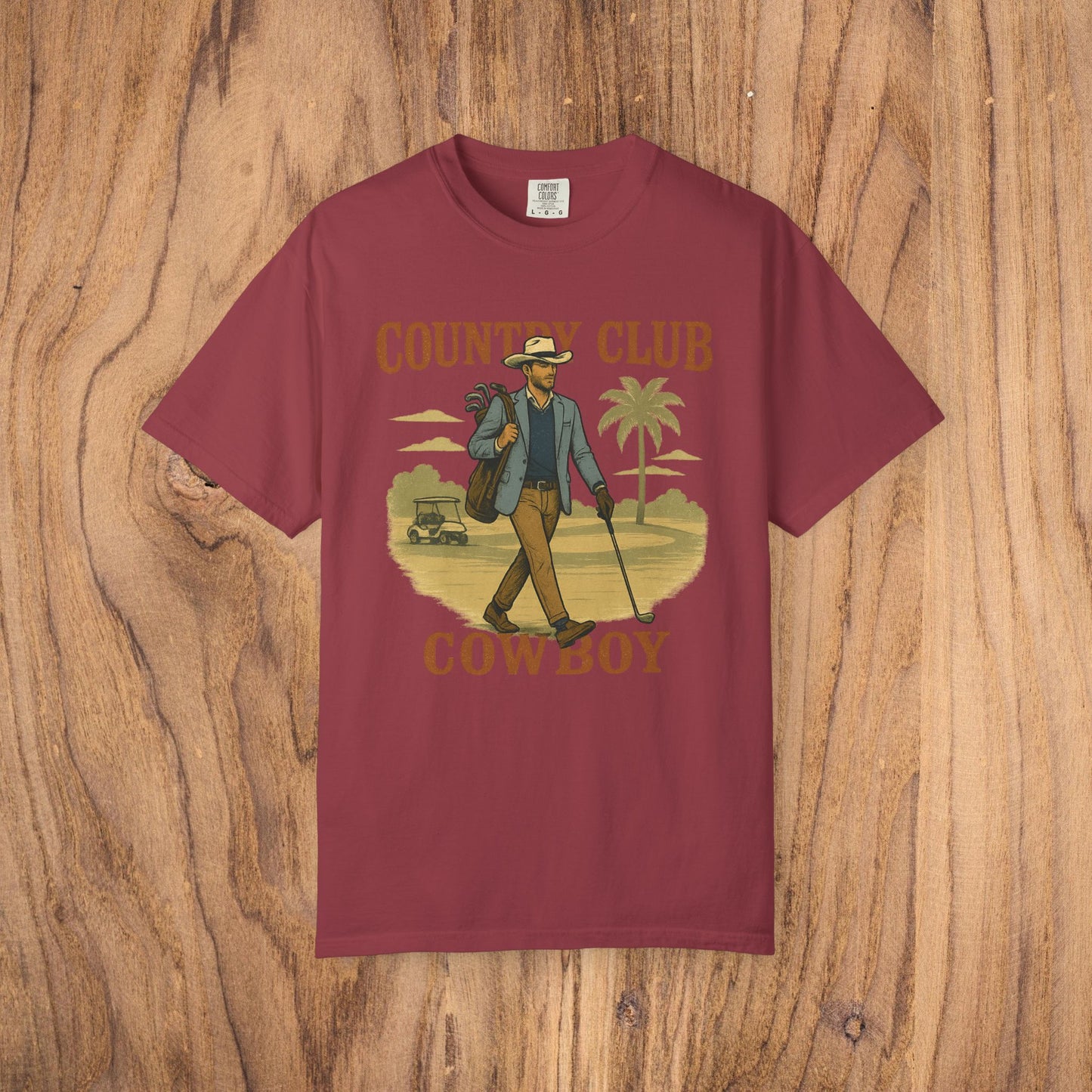 Country Club Cowboy Graphic Tee in maroon color displaying a cowboy in a relaxed pose with a golf bag, surrounded by palm trees, made from high-quality cotton with a ribbed collar and eco-friendly design.