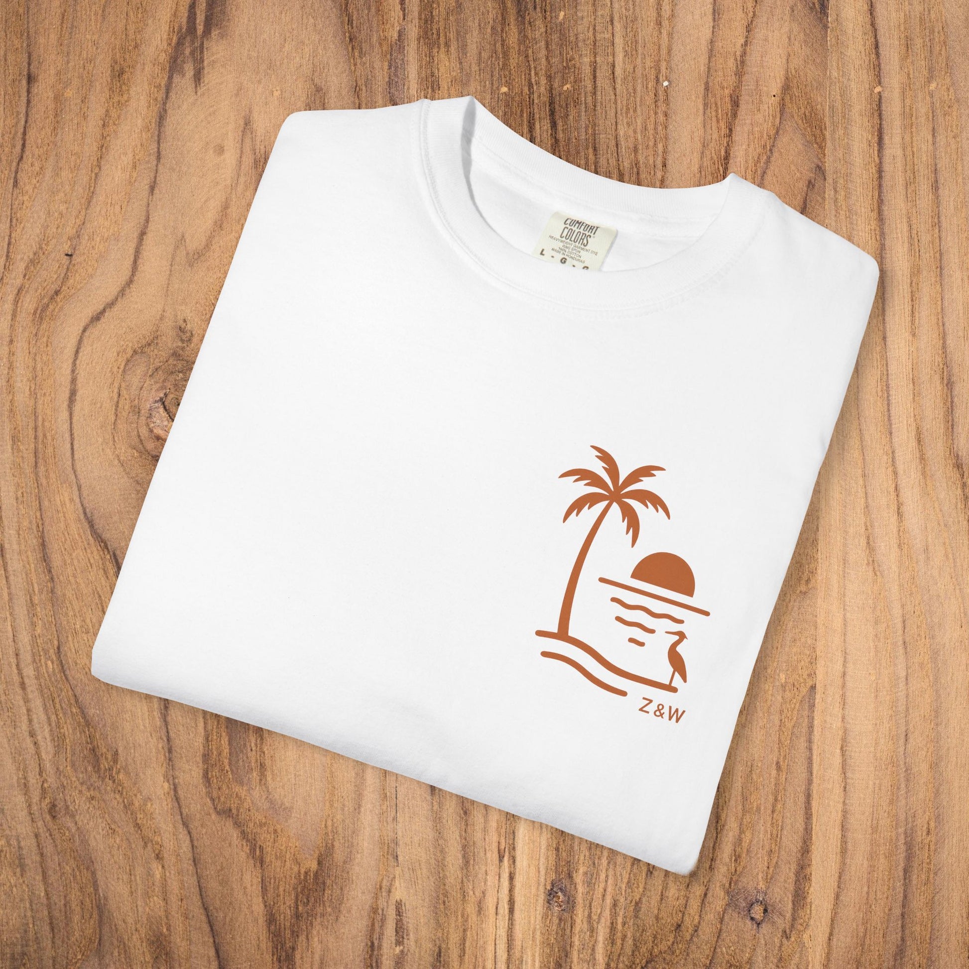 Unisex Sunset Palm Tree T-Shirt displayed flat on a wooden surface, showcasing a vibrant garment-dyed cotton tee in white color, featuring a stylish palm tree and sunset design, perfect for both men and women.