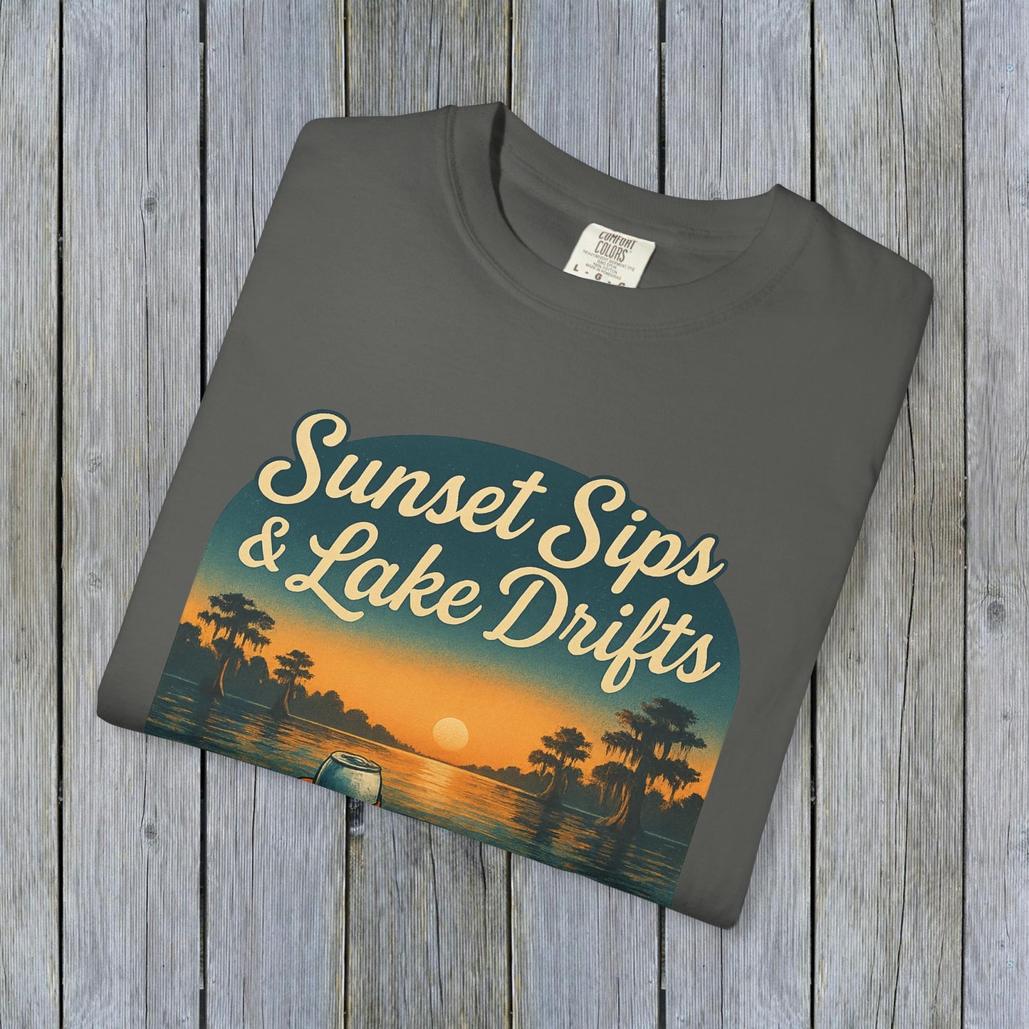 Folded Lake Drifts T-Shirt in soft grey, featuring the captivating 'Sunset Sips & Lake Drifts' graphic on a wooden backdrop.