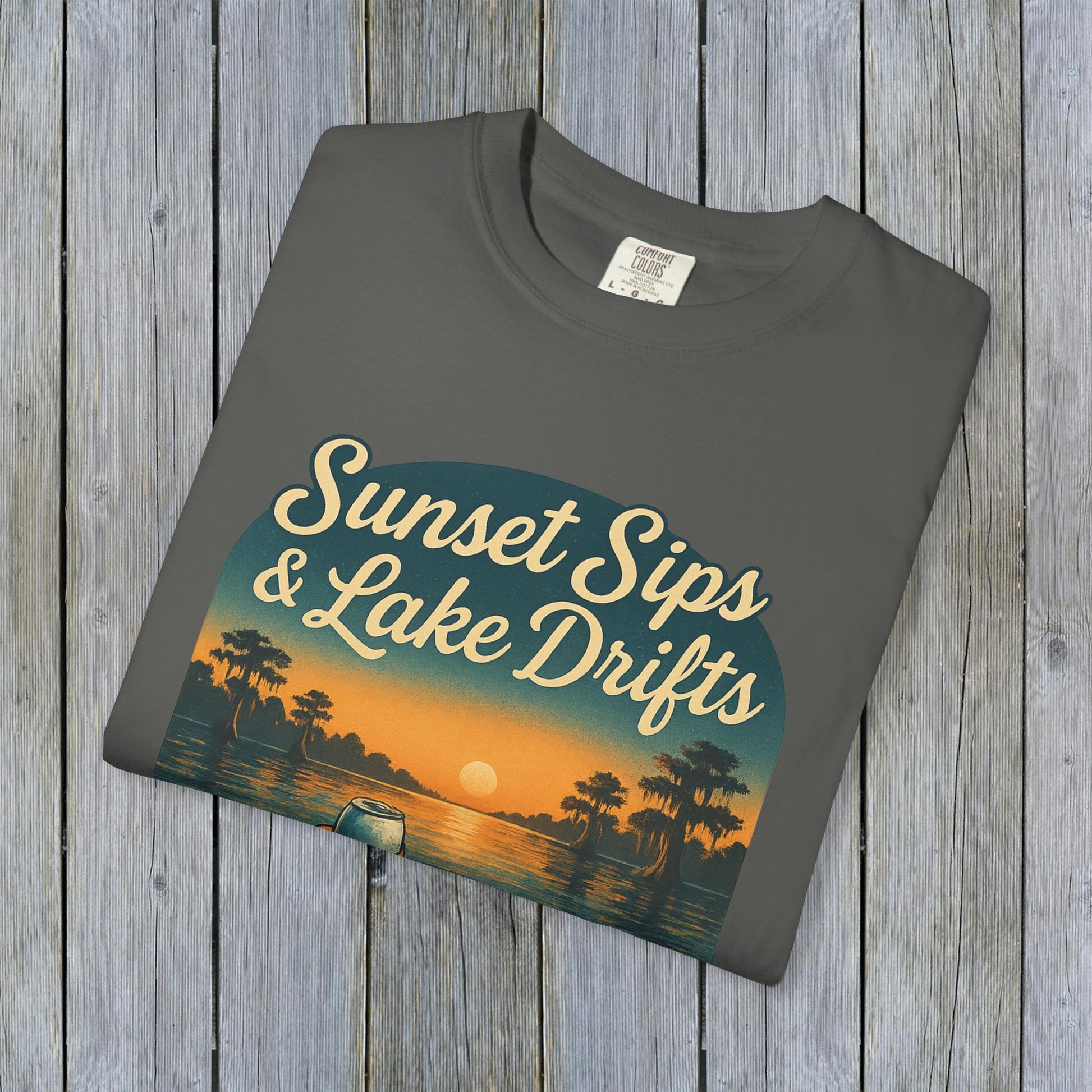 Folded Lake Drifts T-Shirt in soft grey, featuring the captivating 'Sunset Sips & Lake Drifts' graphic on a wooden backdrop.